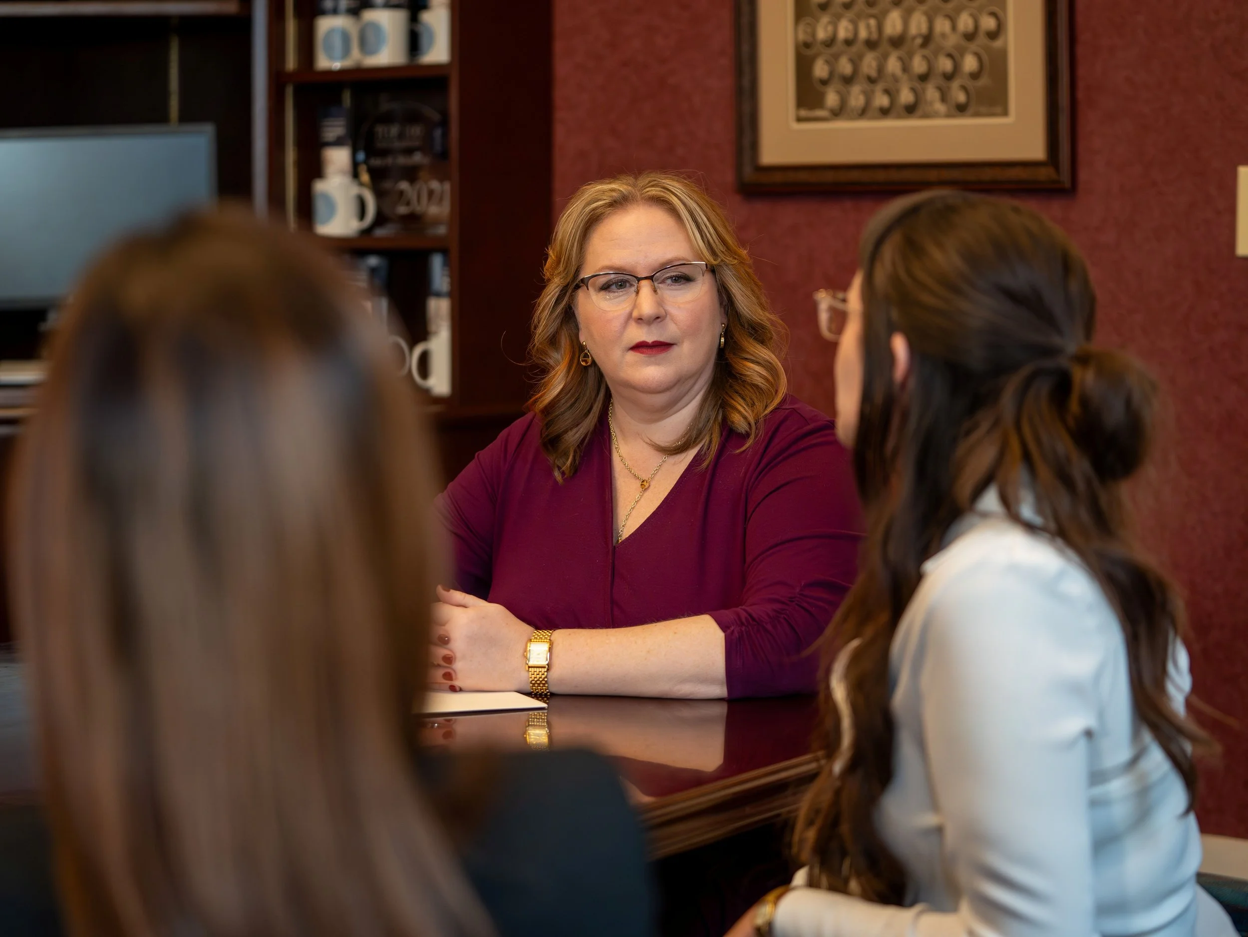 A woman with shoulder-length red hair, glasses, and wearing a burgundy top is engaged in conversation with three women who have their backs to the camera. They are in an office or conference room with a wooden cabinet, framed picture, and a computer in the background.