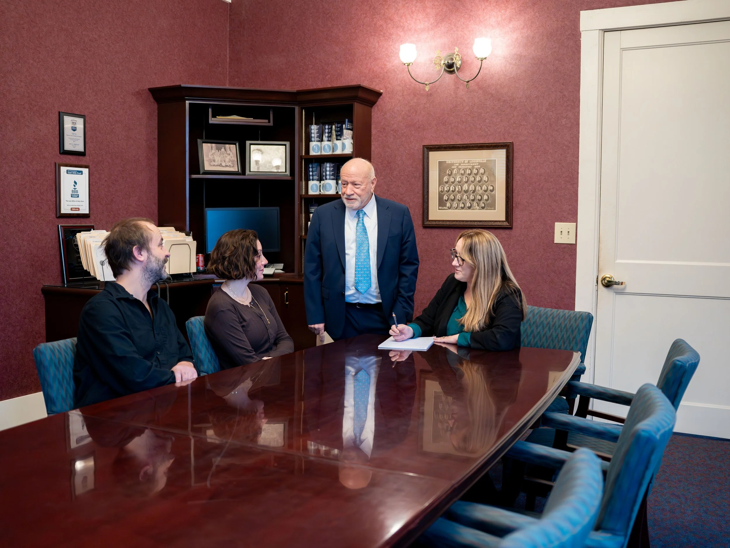 A business meeting in a conference room with four people. Three are seated at a large wooden table, and one man, dressed in a blue suit, is standing. A woman in glasses and a black blazer is writing on a notepad. The room has burgundy walls, framed pictures, and a wooden cabinet behind them.