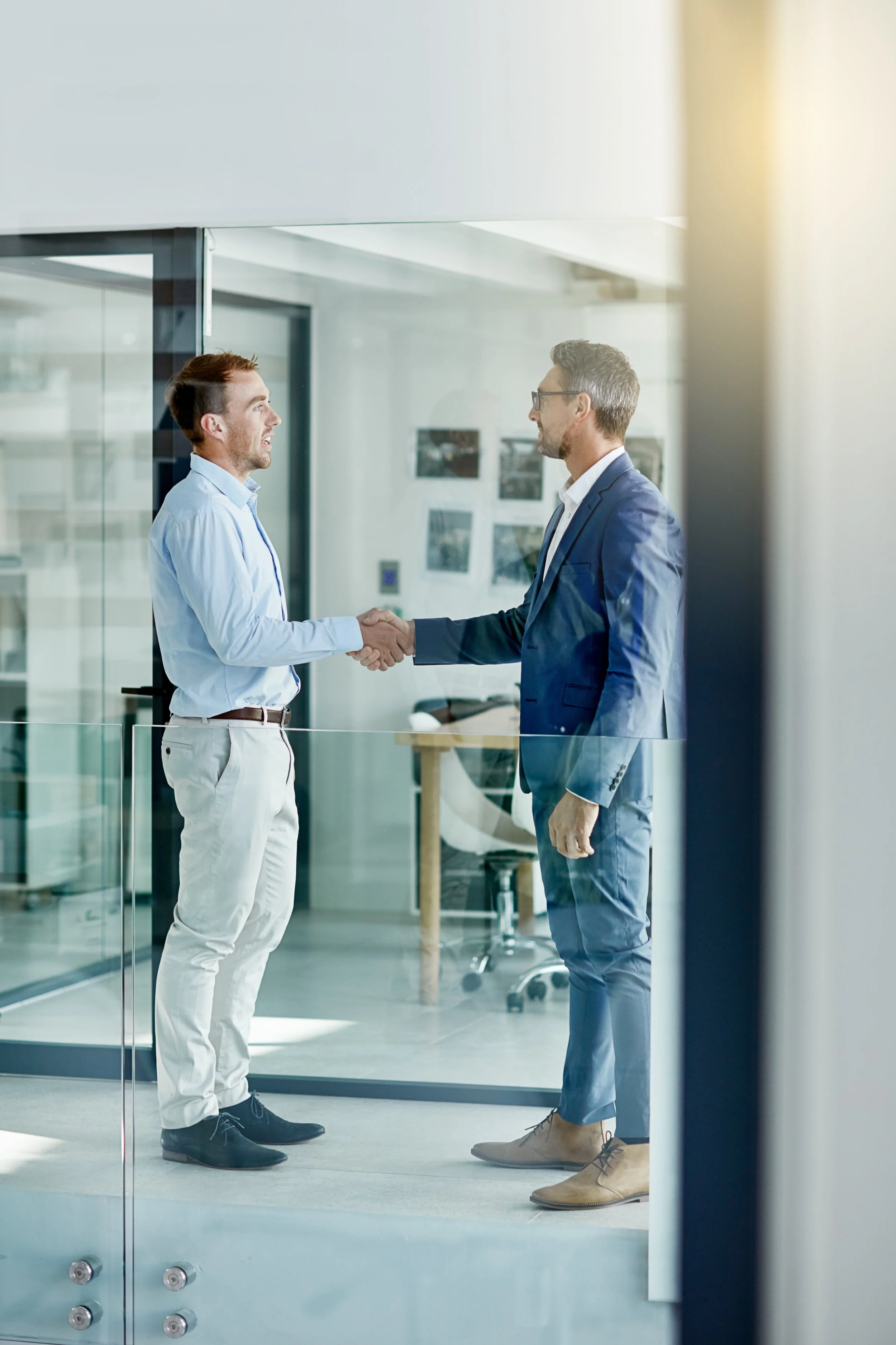 Two men shaking hands in an office setting, one wearing a light blue shirt and white pants, the other in a blue suit and jeans.