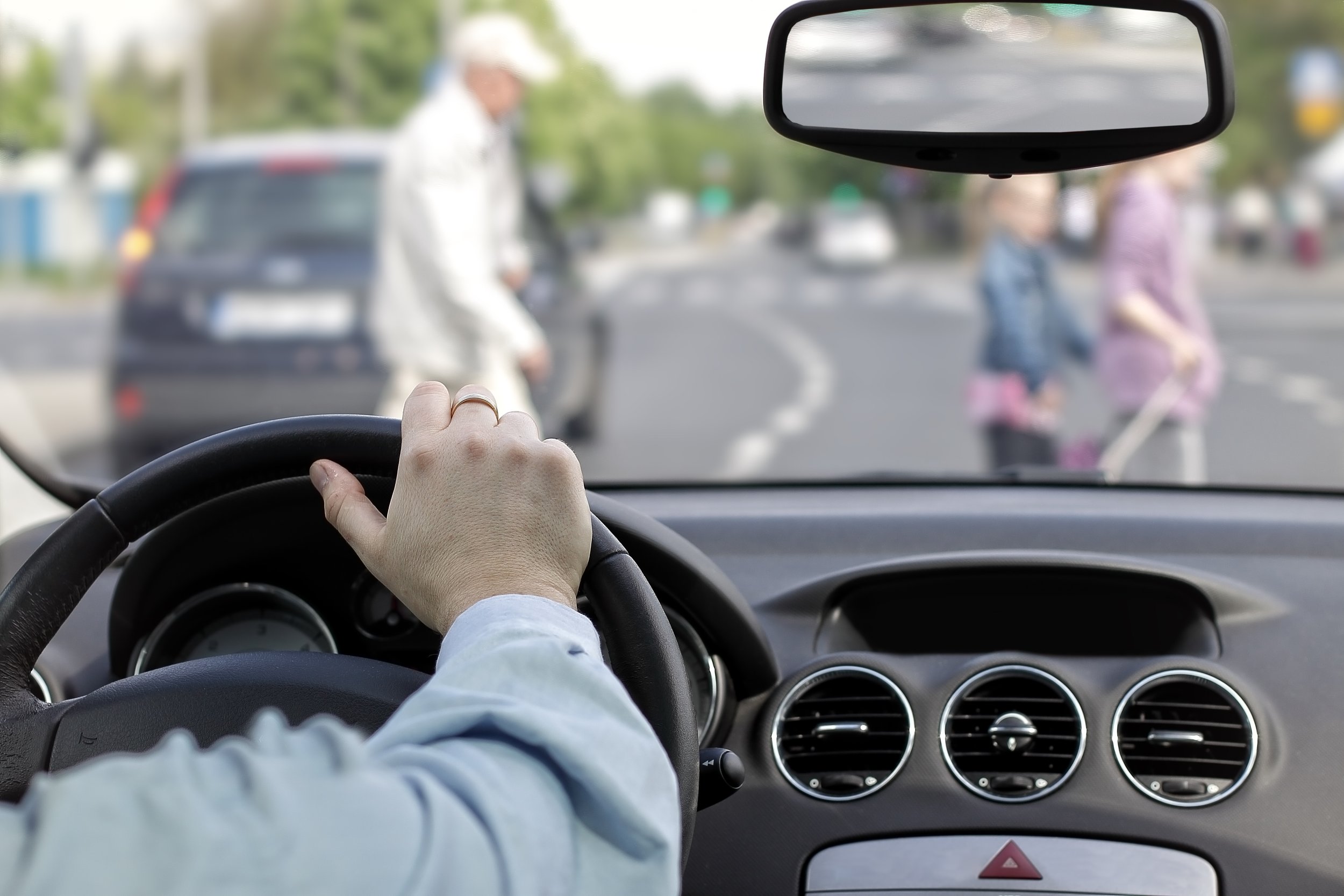 View from inside a car showing the steering wheel, dashboard, and rearview mirror. The car is stopped, and pedestrians are crossing the street in front.
