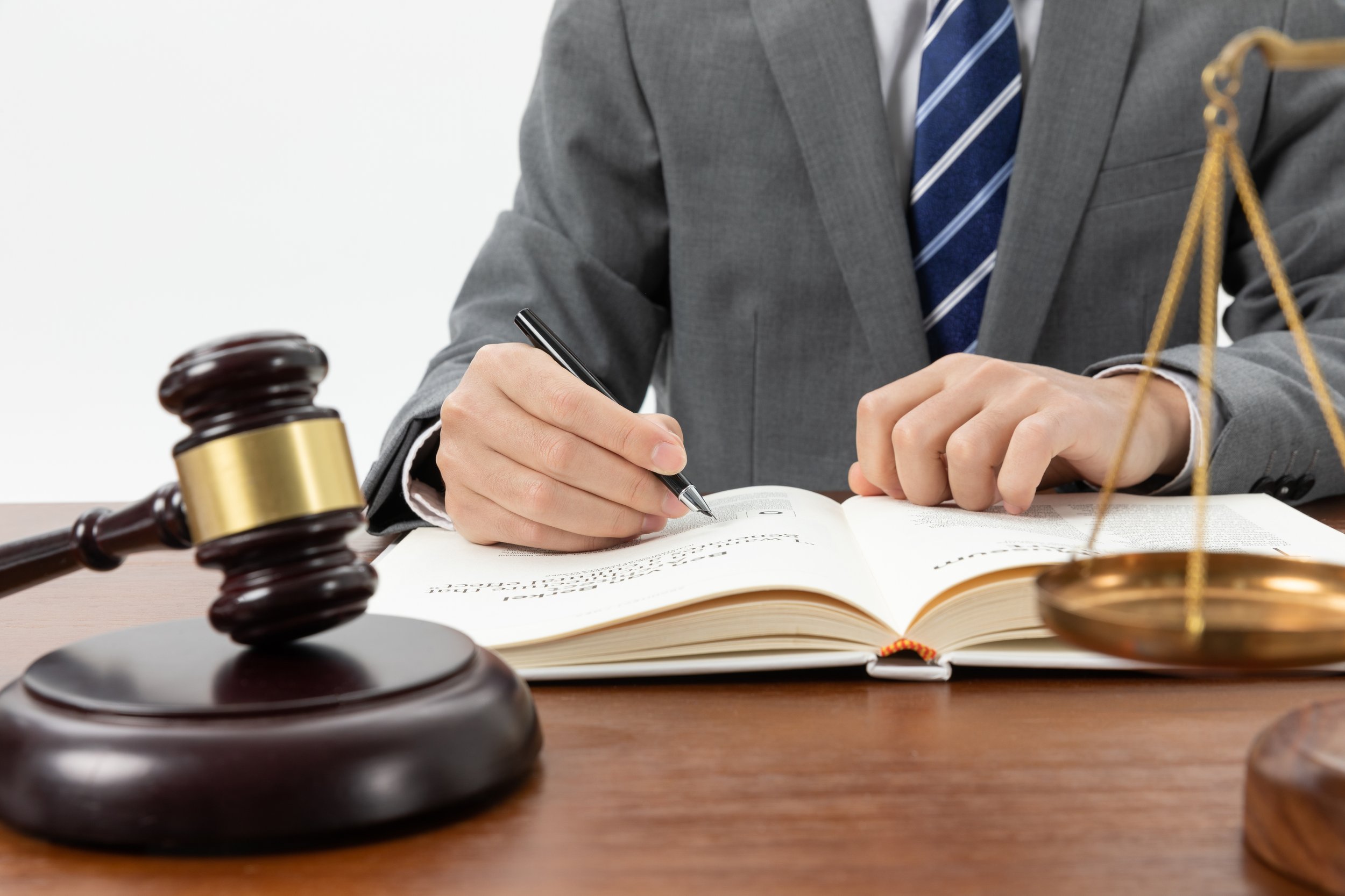 A person in a gray suit and tie sitting at a wooden desk, signing a book, with a gavel and scales of justice on the desk.