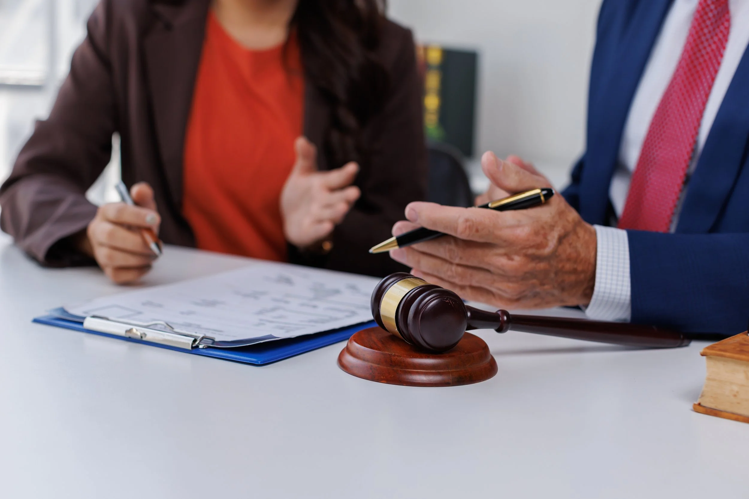A person in a suit holding a pen and discussing legal matters with a woman at a desk with a gavel, a microphone, and documents.