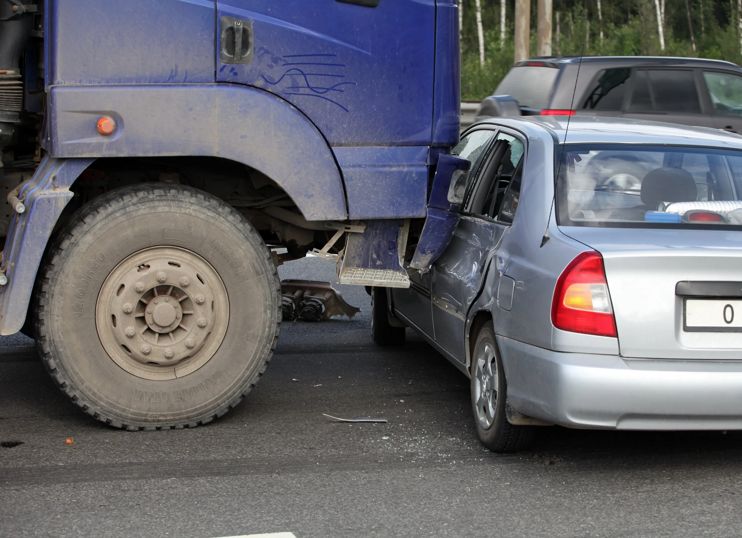 A silver car involved in a collision with a large blue truck, with visible damage to the car's driver side door.