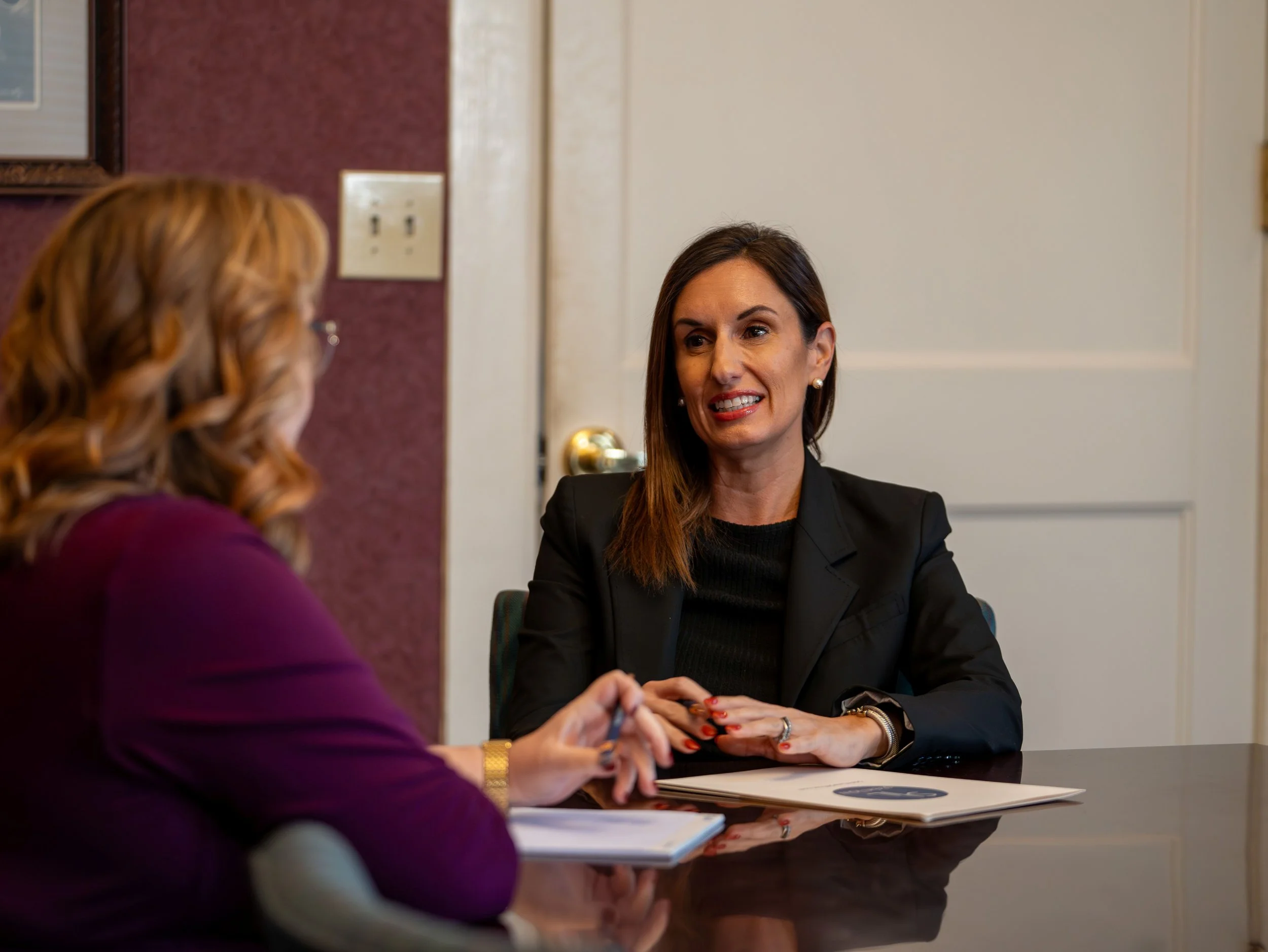 Two women having a discussion at a table in a professional setting. One woman is in focus, wearing a black blazer, and the other woman is blurred in the foreground wearing a purple top.