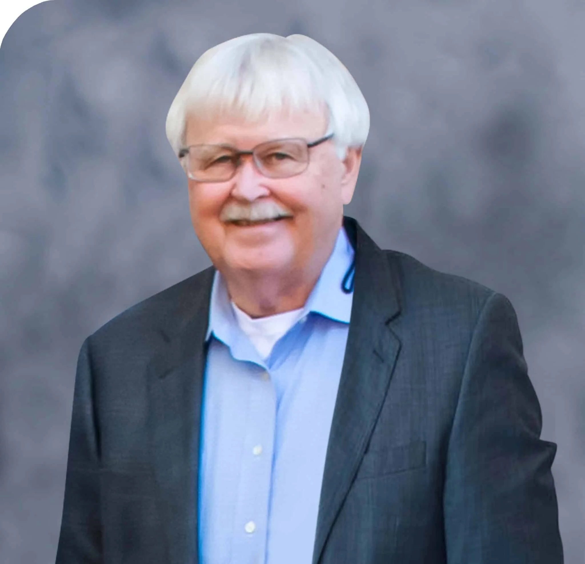 A middle-aged man with white hair and glasses, smiling, dressed in a suit jacket over a light blue shirt, standing outdoors with a blurred cloudy sky background.