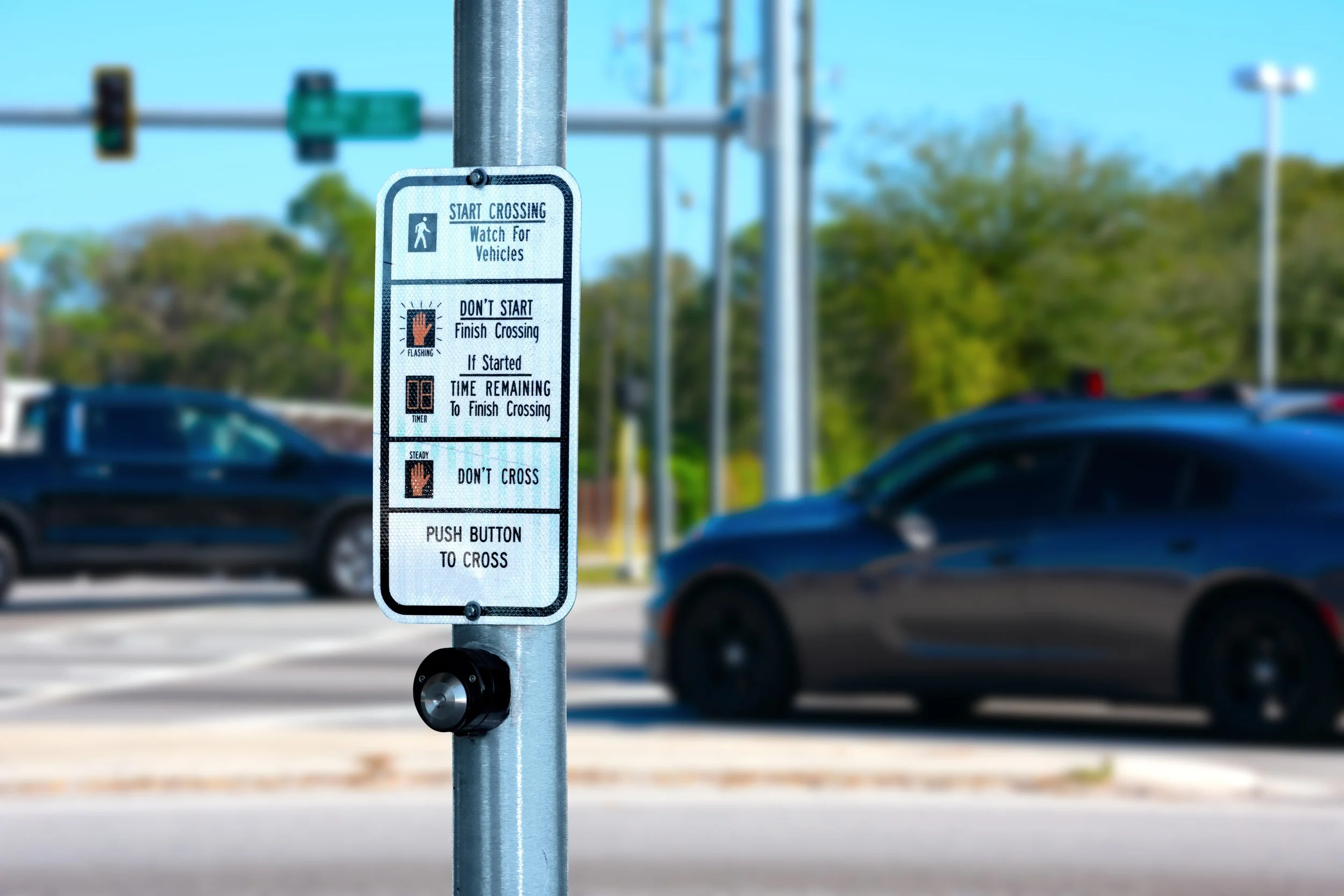 Traffic crossing instructions sign on a pole, with cars in the background, instructing pedestrians to watch for vehicles, not start crossing if the red flashing light is on, and to push a button to cross.