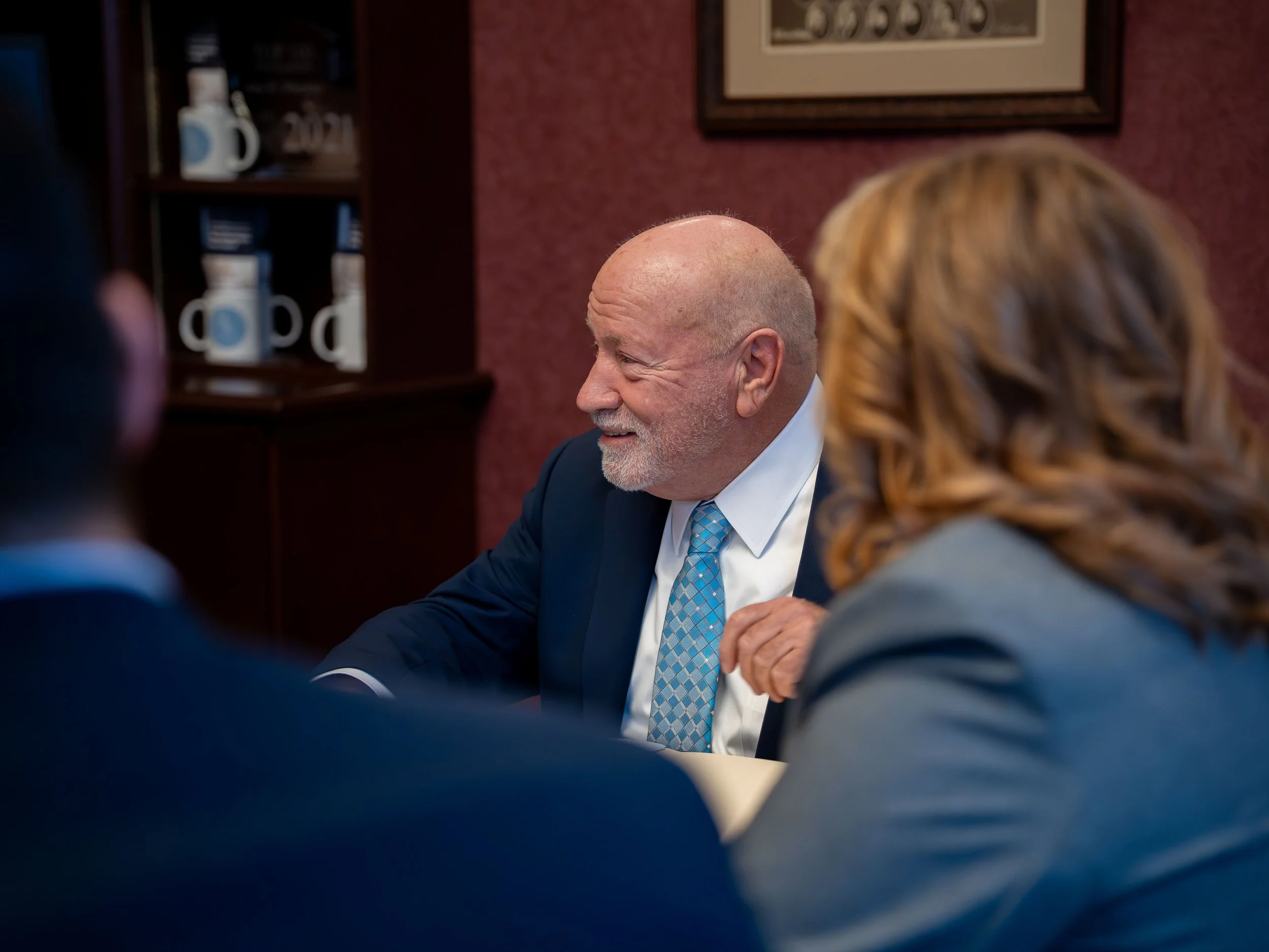 A smiling man with a bald head and gray beard wearing a dark suit and a blue patterned tie, sitting at a table with a woman with long wavy red hair, in a room with a bookshelf and framed art on the wall.