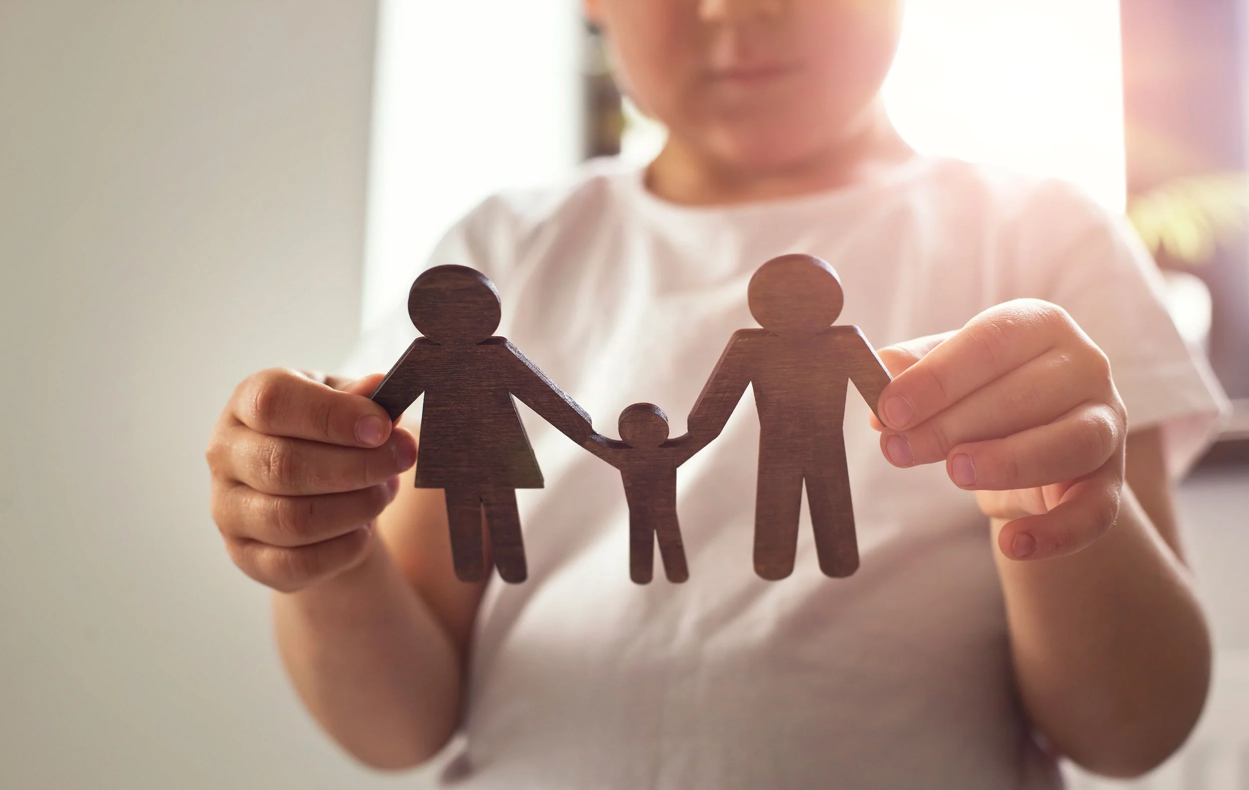 Child holding wooden cutouts of a family of three, with the child in the center holding hands with two larger figures representing a family.