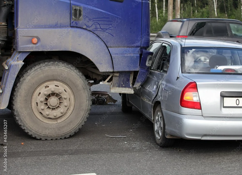 A large blue truck has collided with the side of a silver car, causing significant damage to the car's door and side panels.