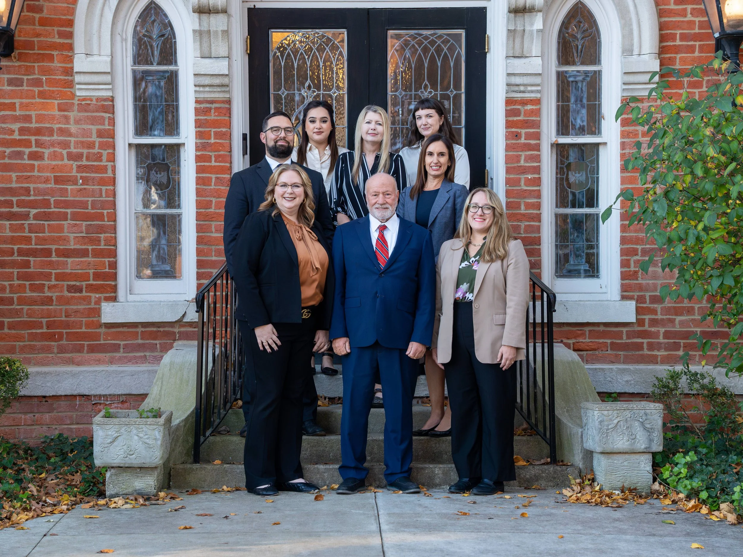 Group of nine professionally dressed people standing on church steps in front of a brick building with stained glass windows, smiling and posing for a photo.
