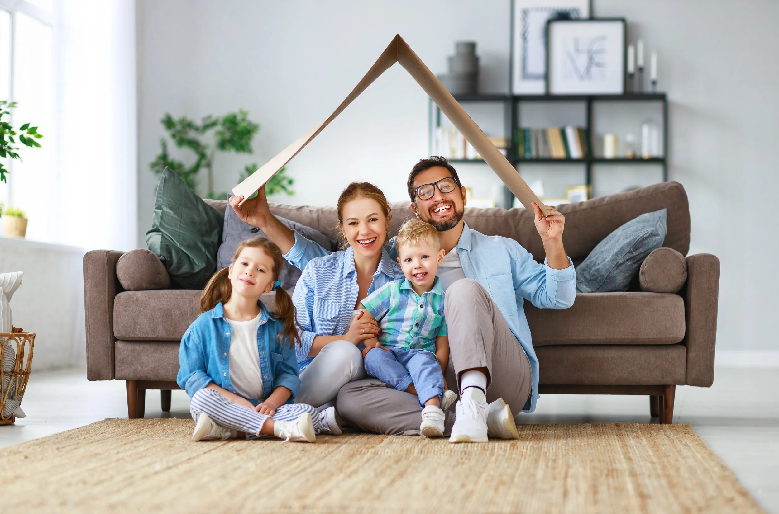 A family of four sitting on the floor in front of a sofa, smiling at the camera. The father and mother are holding a large cardboard piece above their heads to form a house-shaped outline. Two children, a girl and a boy, sit in front of them. The background shows a living room with a bookshelf and some framed pictures.