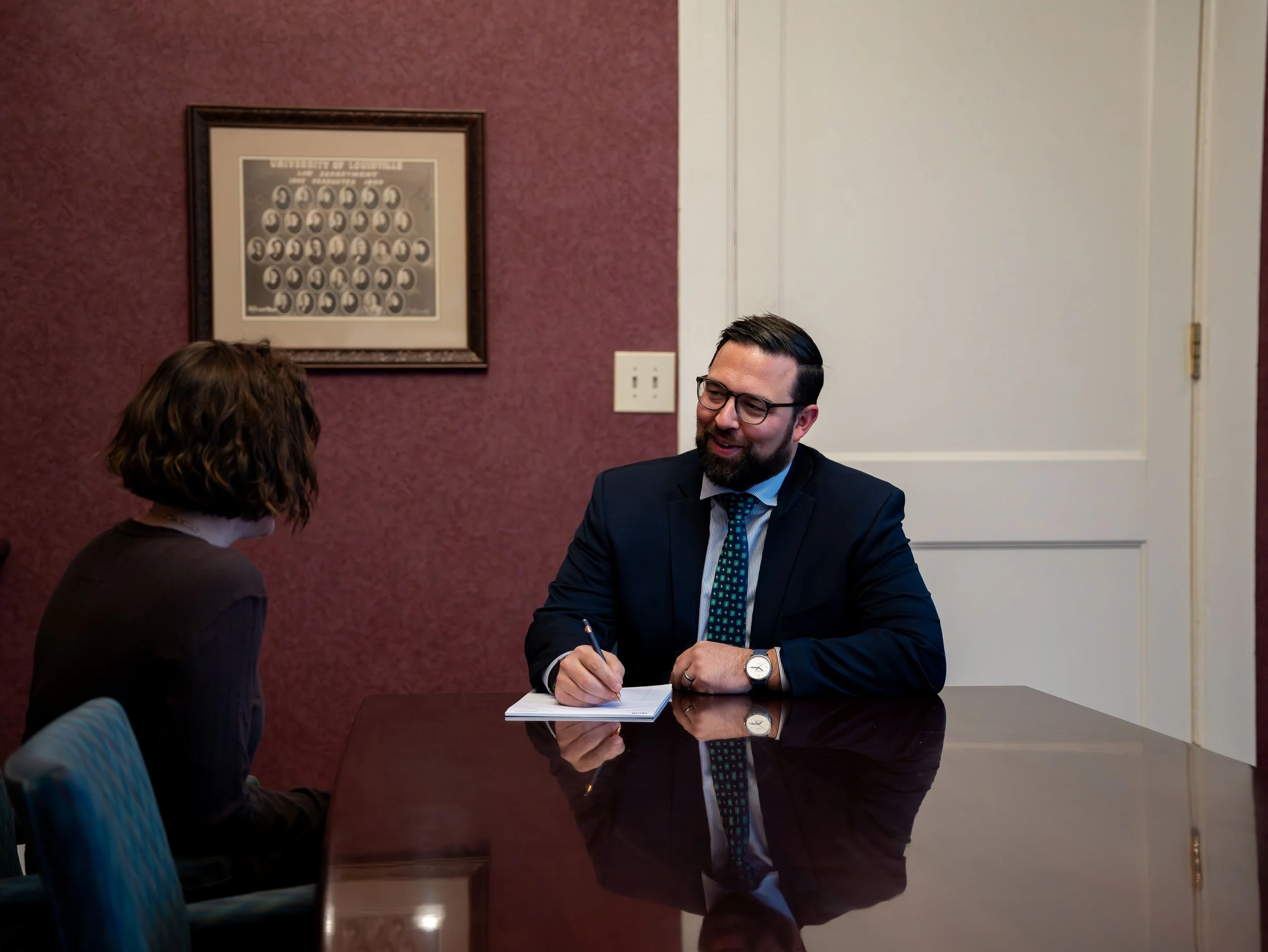 A man in a suit and glasses sitting at a table, talking to a woman with shoulder-length hair, in an office setting.