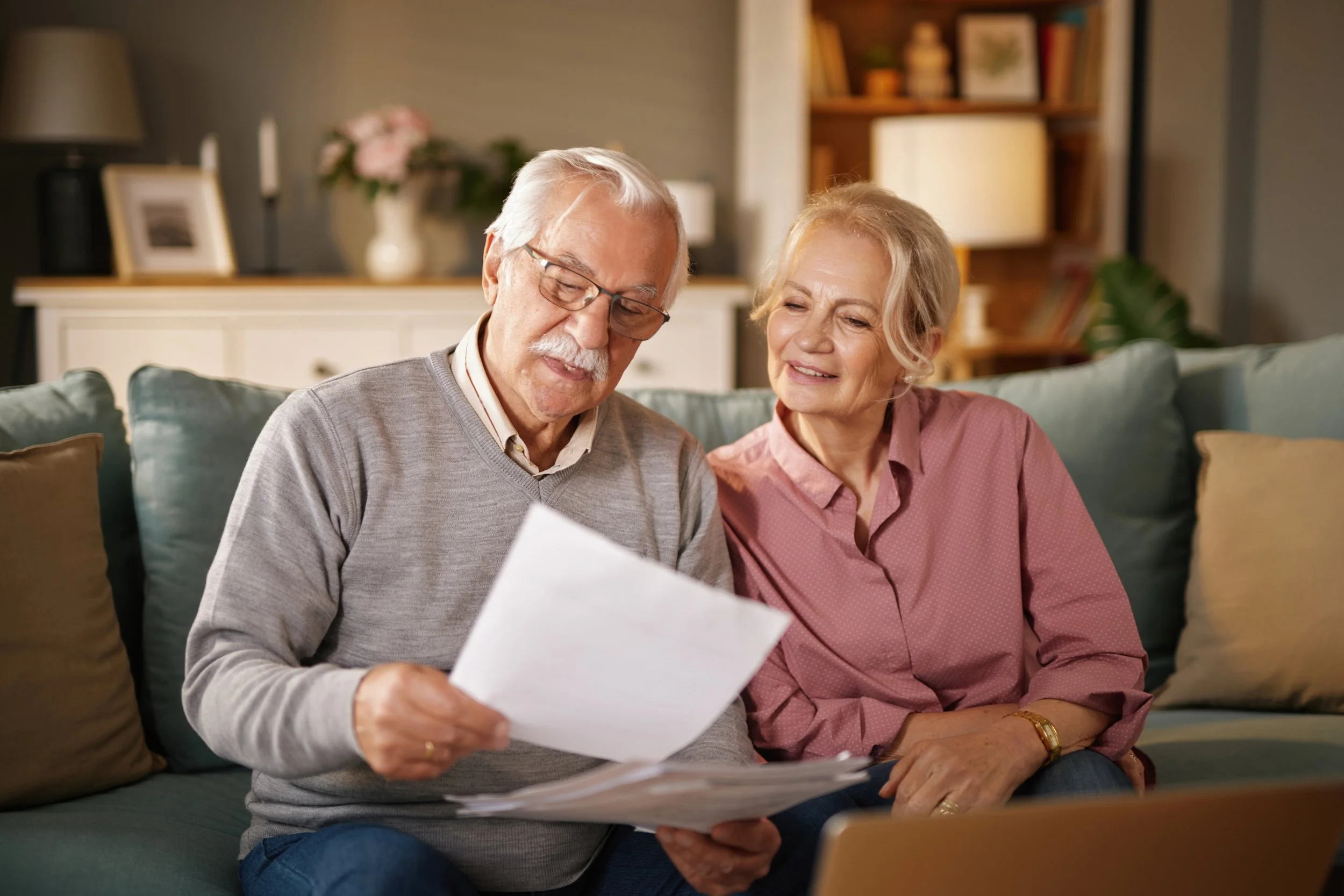 An elderly couple sitting on a couch, looking over papers and documents, smiling and discussing together in a cozy living room.