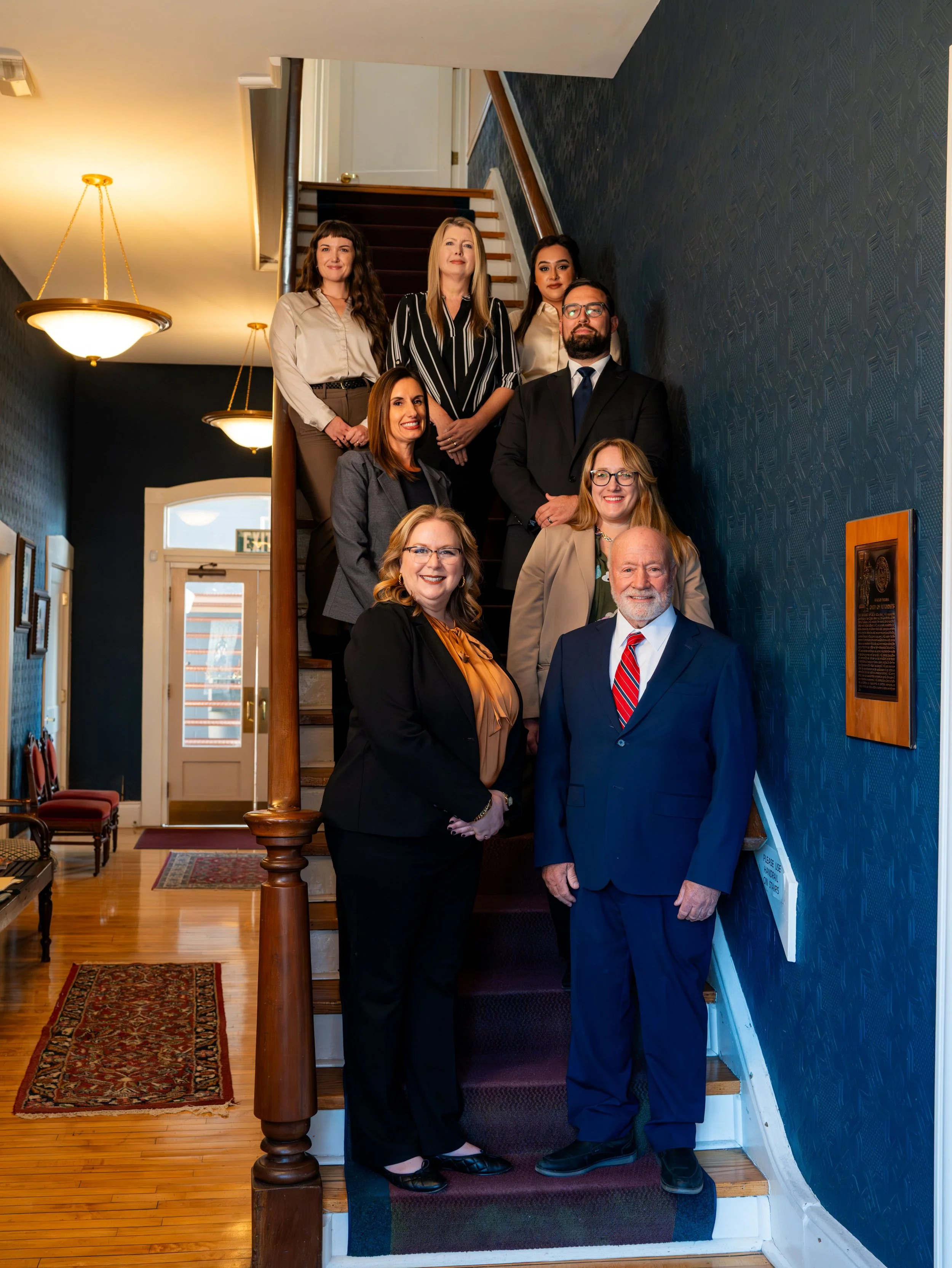 Group of nine professionals dressed in business attire standing on and around a staircase in a hallway of a building.