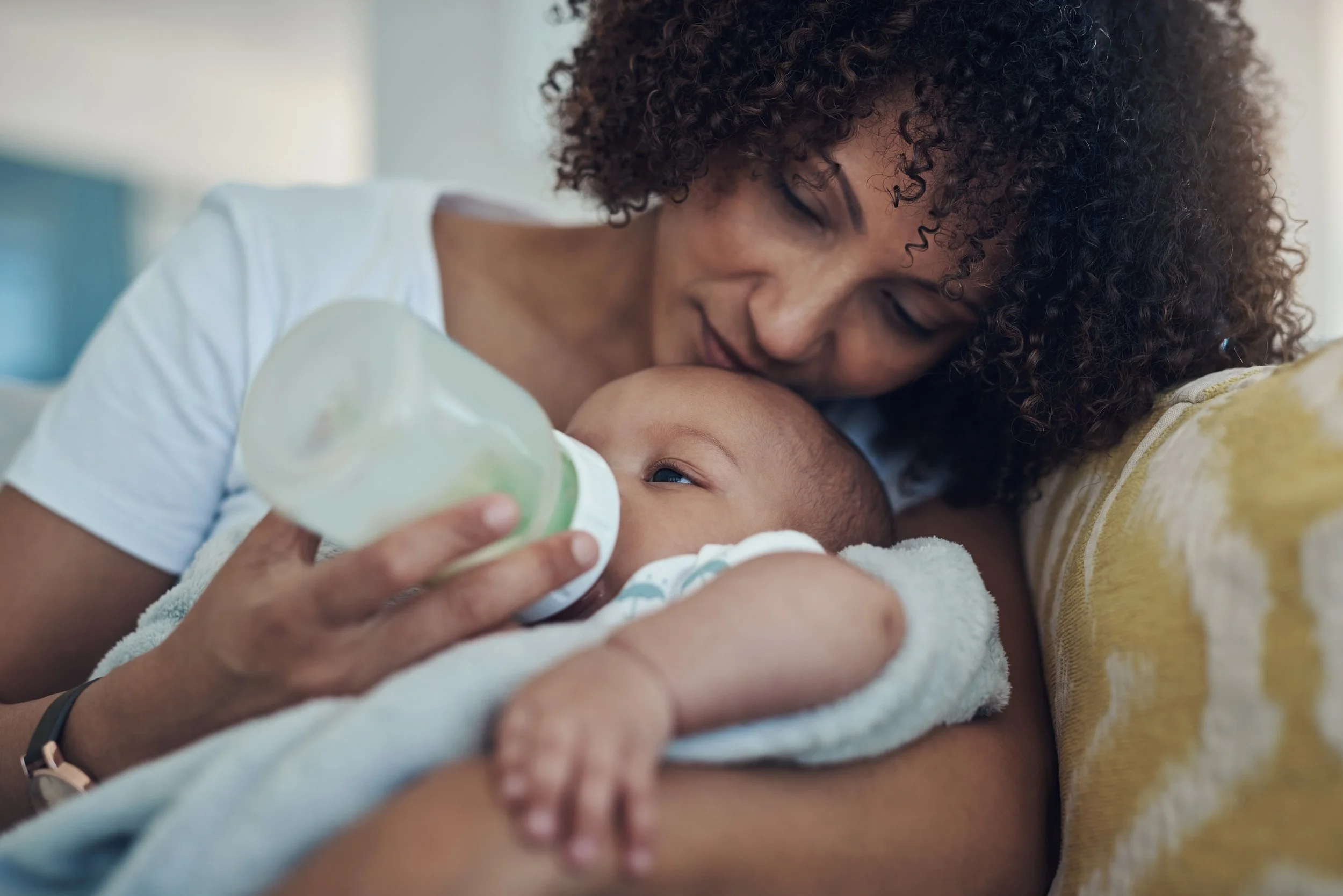 Close-up of a woman with curly hair smiling gently as she feeds a baby with a bottle, lying on a blanket.