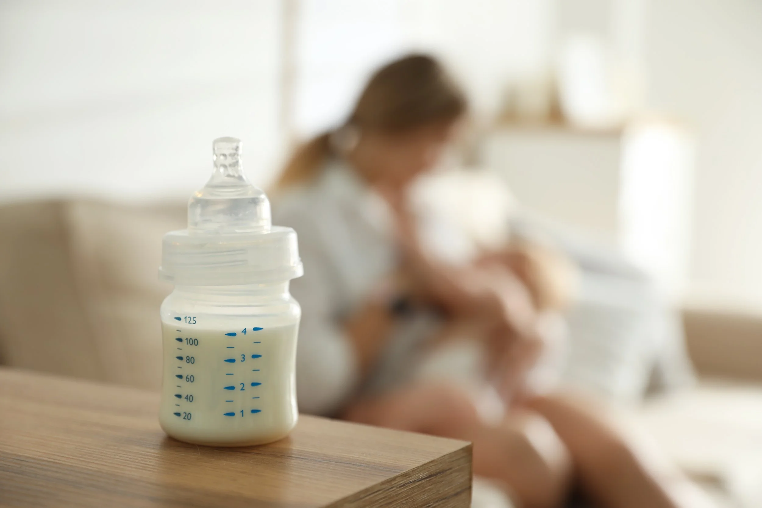 A close-up of a baby bottle filled with milk placed on a wooden surface, with a blurred woman sitting in the background.