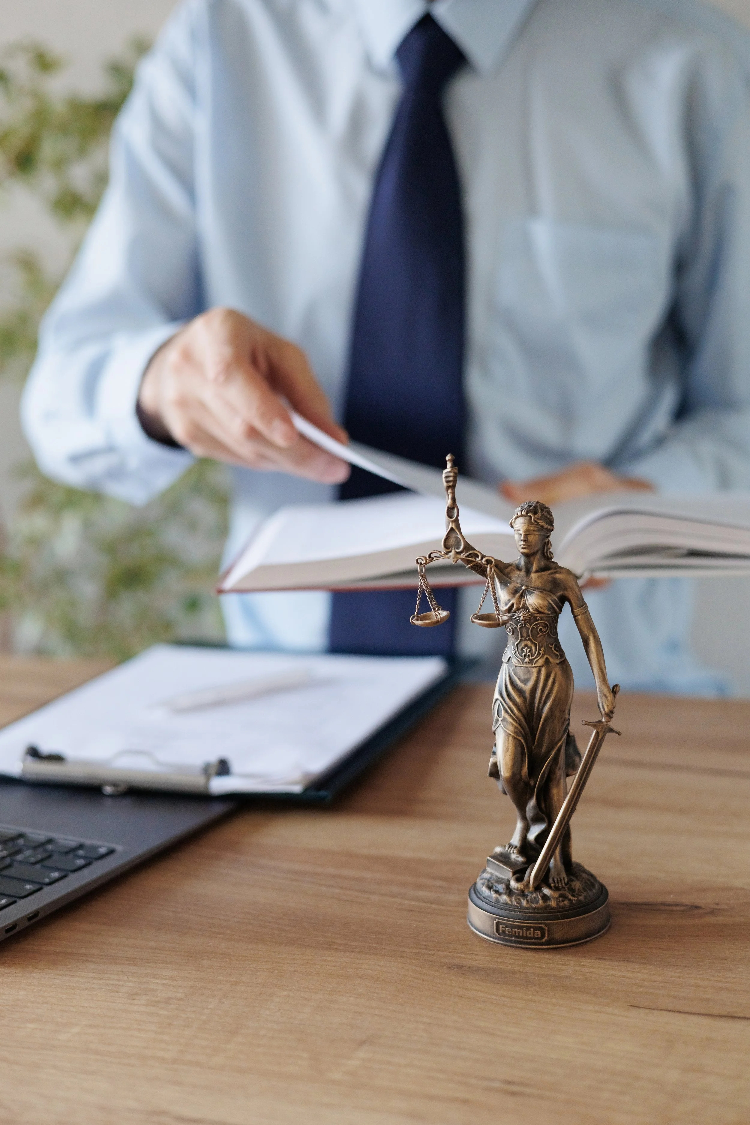 Close-up of a lawyer's desk with a bronze statue of Lady Justice, a laptop, and a clipboard, with a lawyer in a light blue shirt and dark tie in the background.