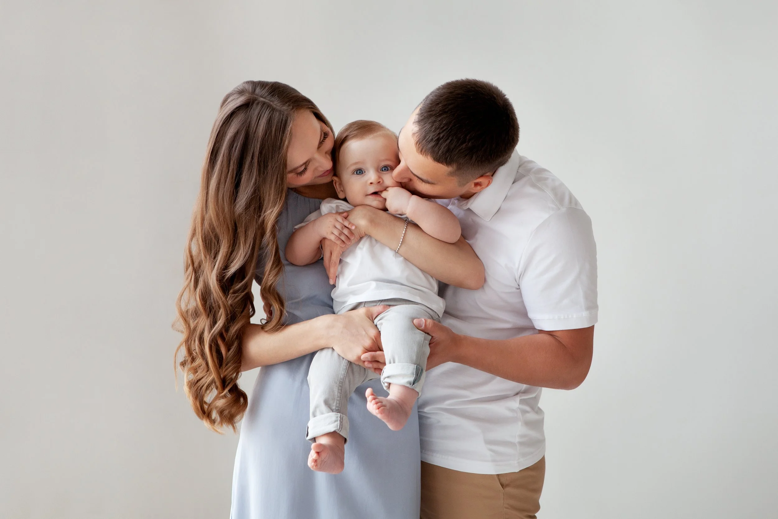 A family of three consists of a woman, a man, and a baby. They are smiling and playing together against a plain, light-colored background. The woman has long, wavy brown hair and is holding the baby, who has blue eyes and light brown hair. The man has short dark hair, is wearing a white shirt, and is kissing the baby's face.