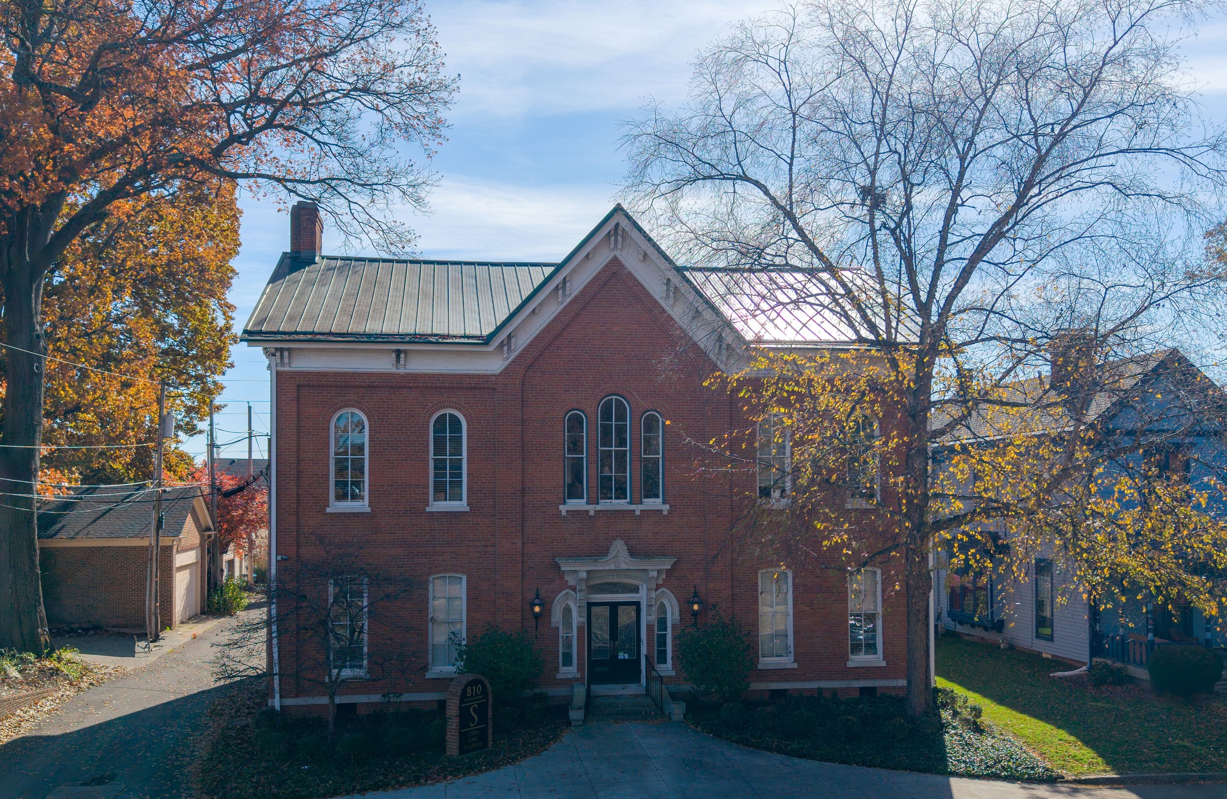 A large red brick house with tall arched windows, a steep roof with a chimney, and an entryway with a small porch and two lamps flanking the door. There are trees with autumn leaves surrounding the house, and a driveway leading up to it.