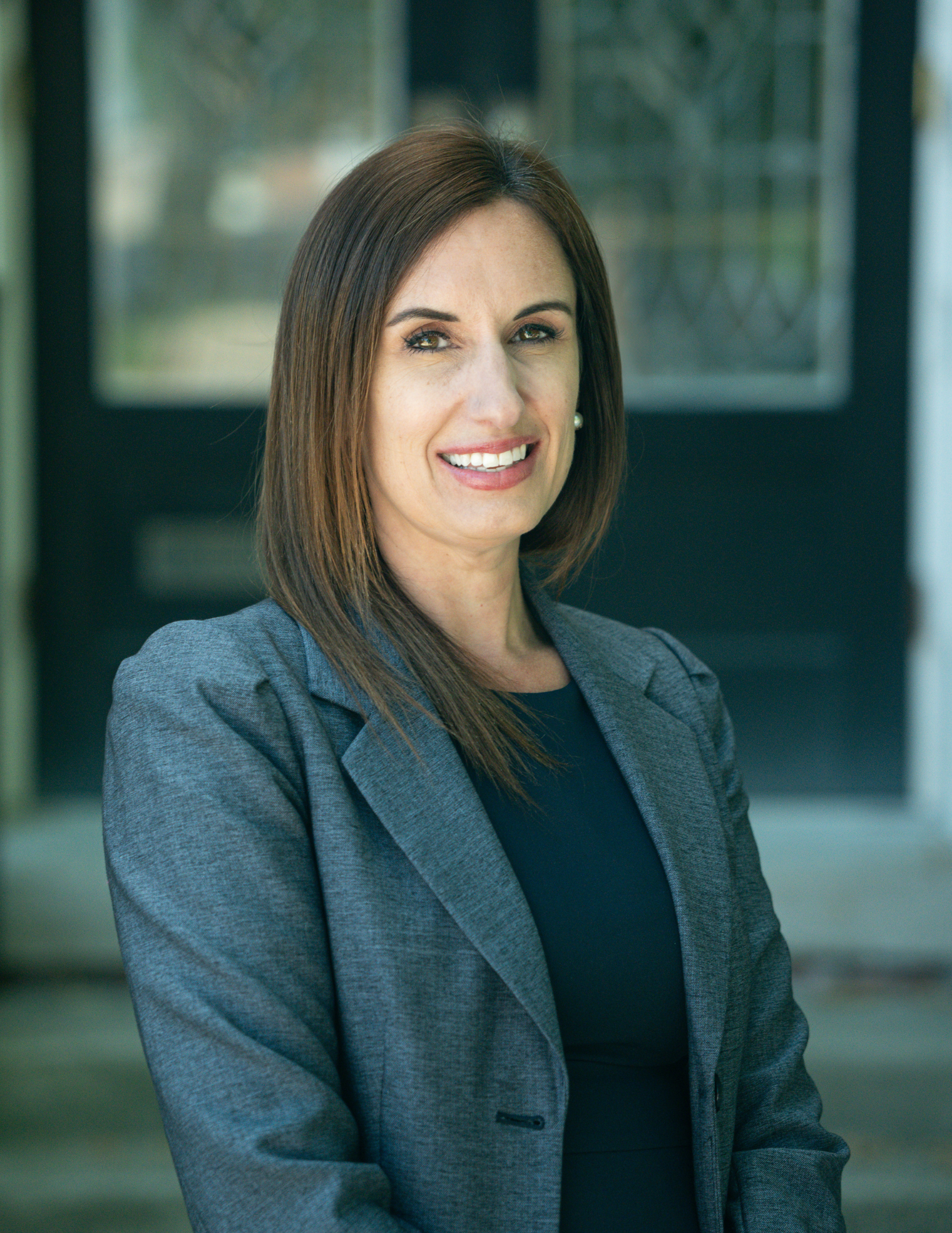 a woman with straight, shoulder-length brown hair, wearing a dark gray blazer and smiling with her arms crossed, standing outdoors near a modern building with glass windows