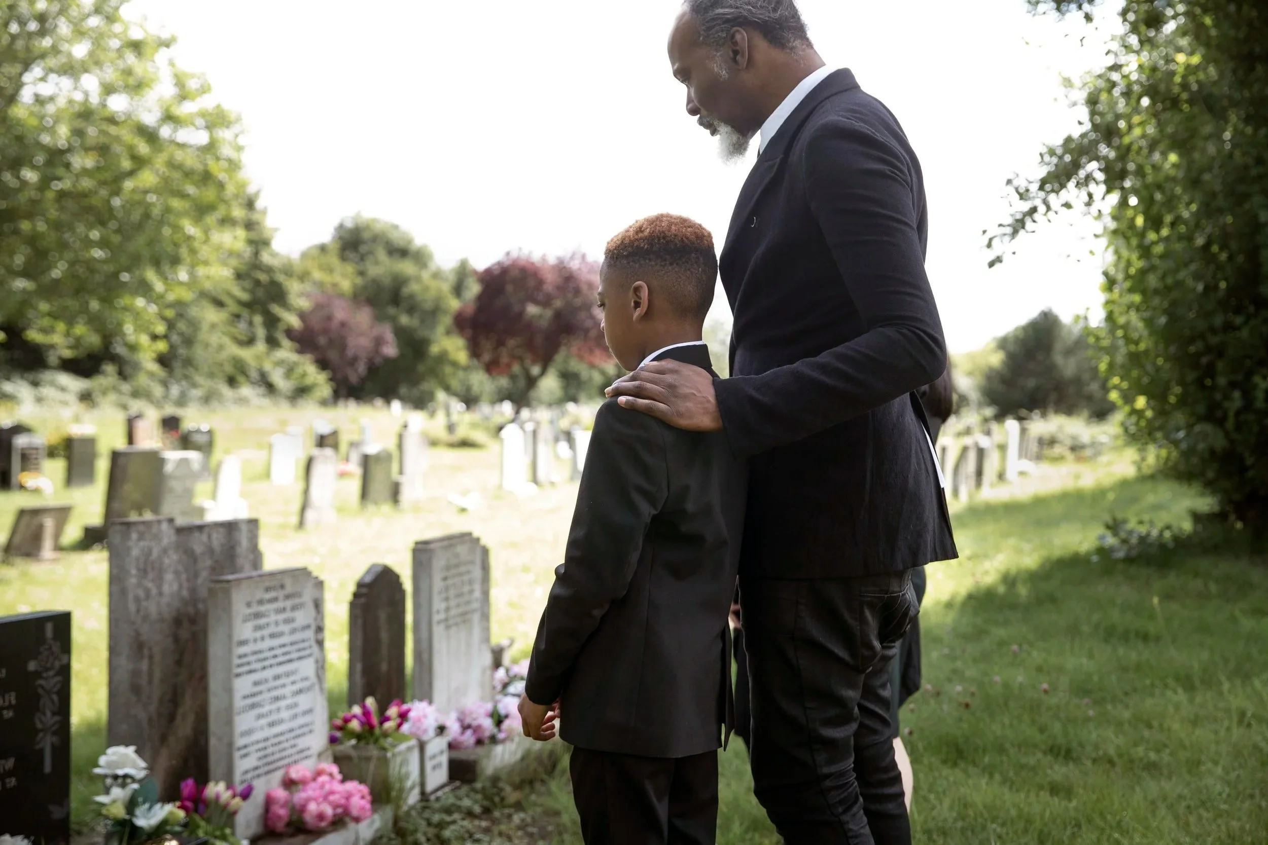 A man and a boy stand together at a cemetery, mourning at a grave with flowers, during daytime.