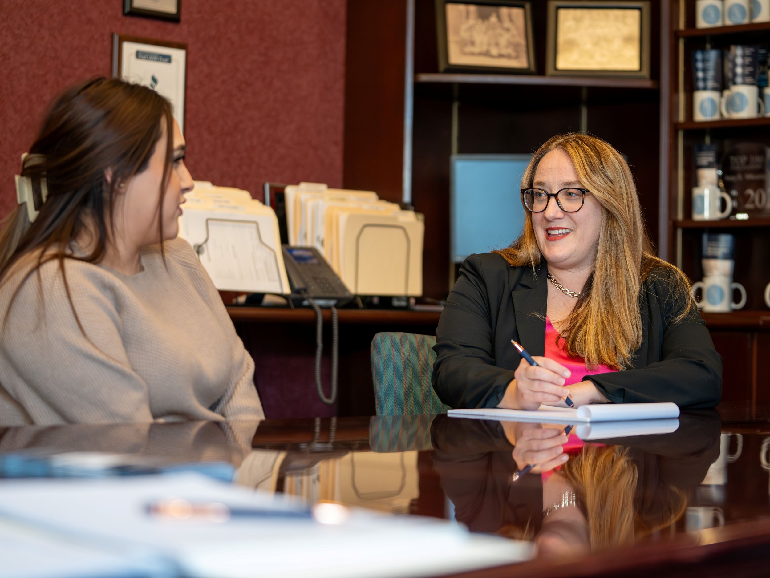 Two women having a conversation at a meeting table in an office or conference room, with documents and office supplies on the table and shelves in the background.