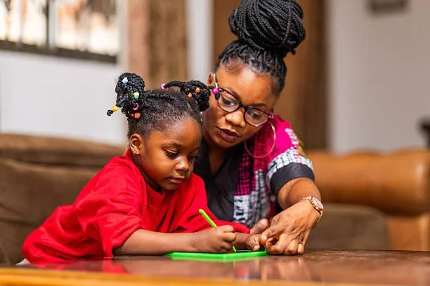 A woman and a young girl with braided hair and colorful beads sit together at a wooden table, engaging in a coloring activity with green crayons. The woman wears glasses and a pink, black, and gray top, while the girl wears a red shirt.