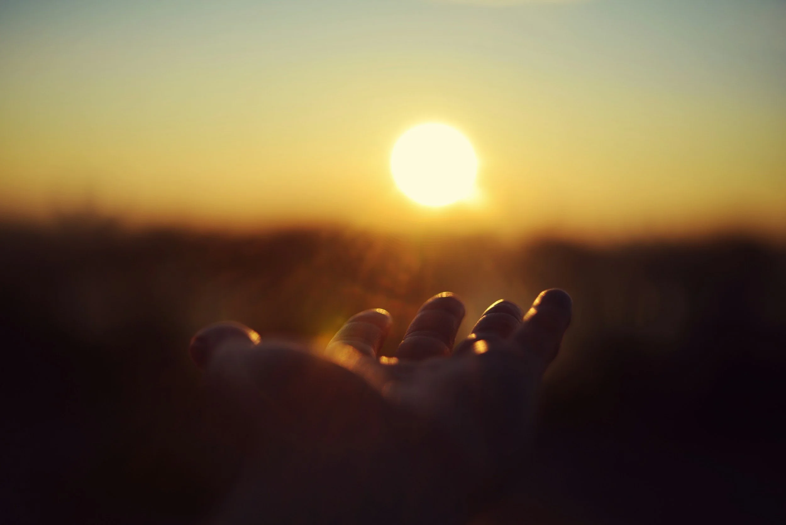 A hand reaching out towards a setting sun on the horizon during sunset, with a blurred landscape in the background.