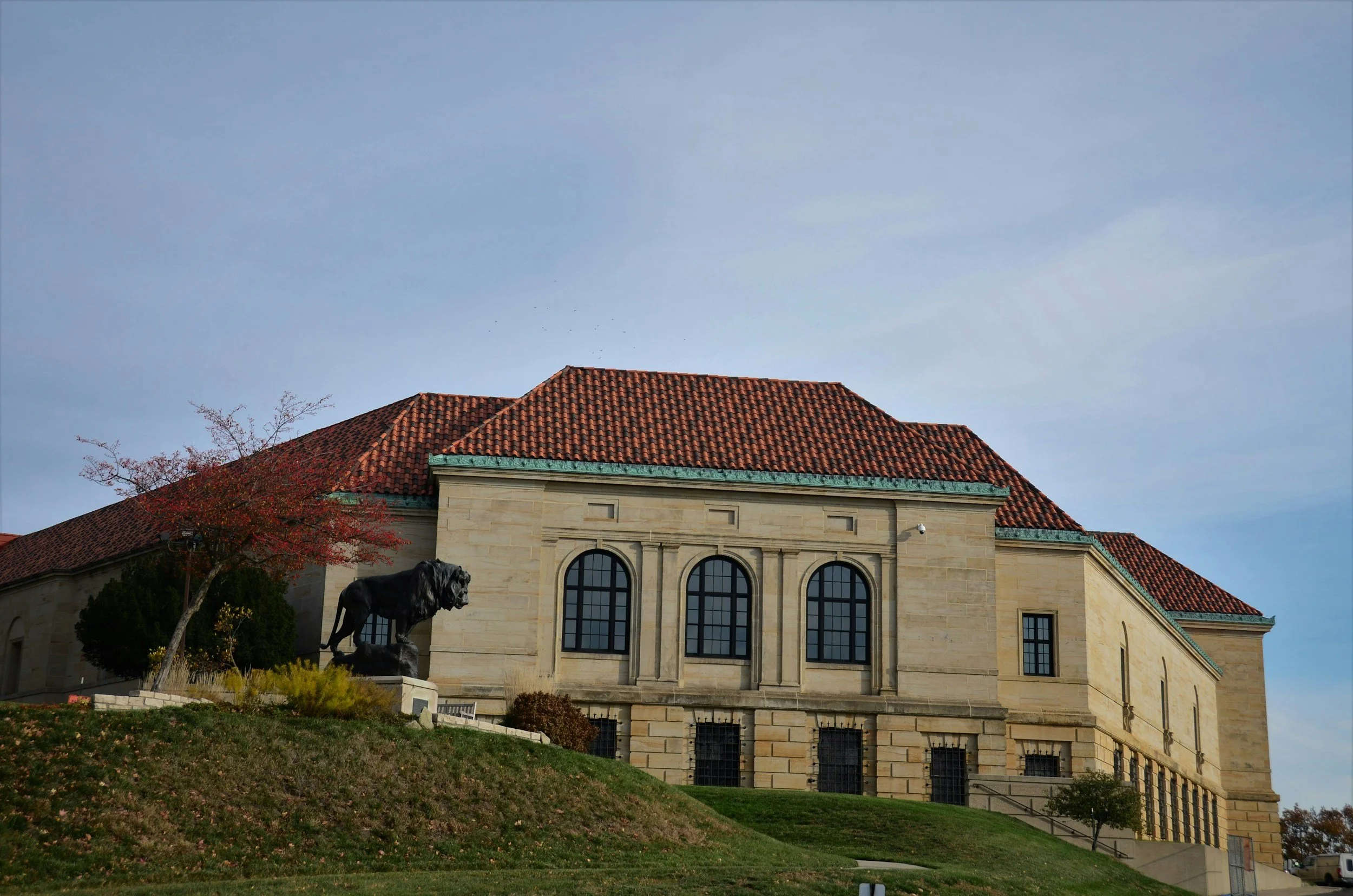 A large historic building with a red tile roof, beige stone exterior, and arched windows, situated on a grassy hill with a black lion statue at its base and a small tree with red leaves nearby.