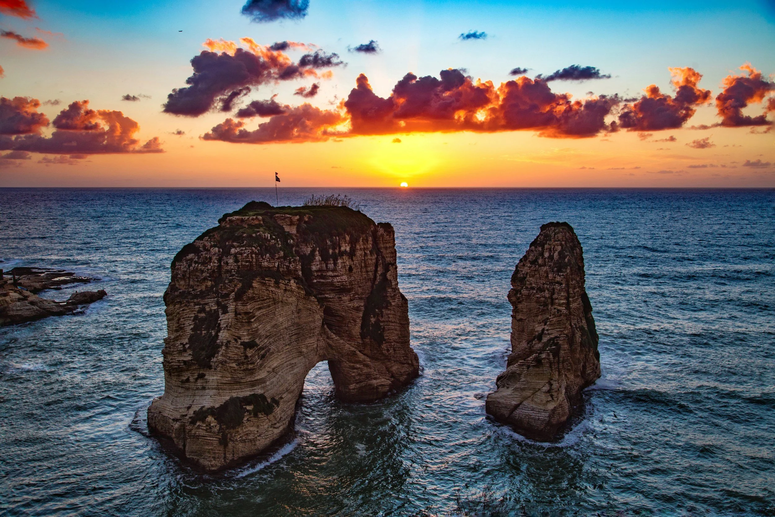 Sunset over the ocean with two large rock formations in the water, one of which has an arch, and a small flag on top of the left formation.