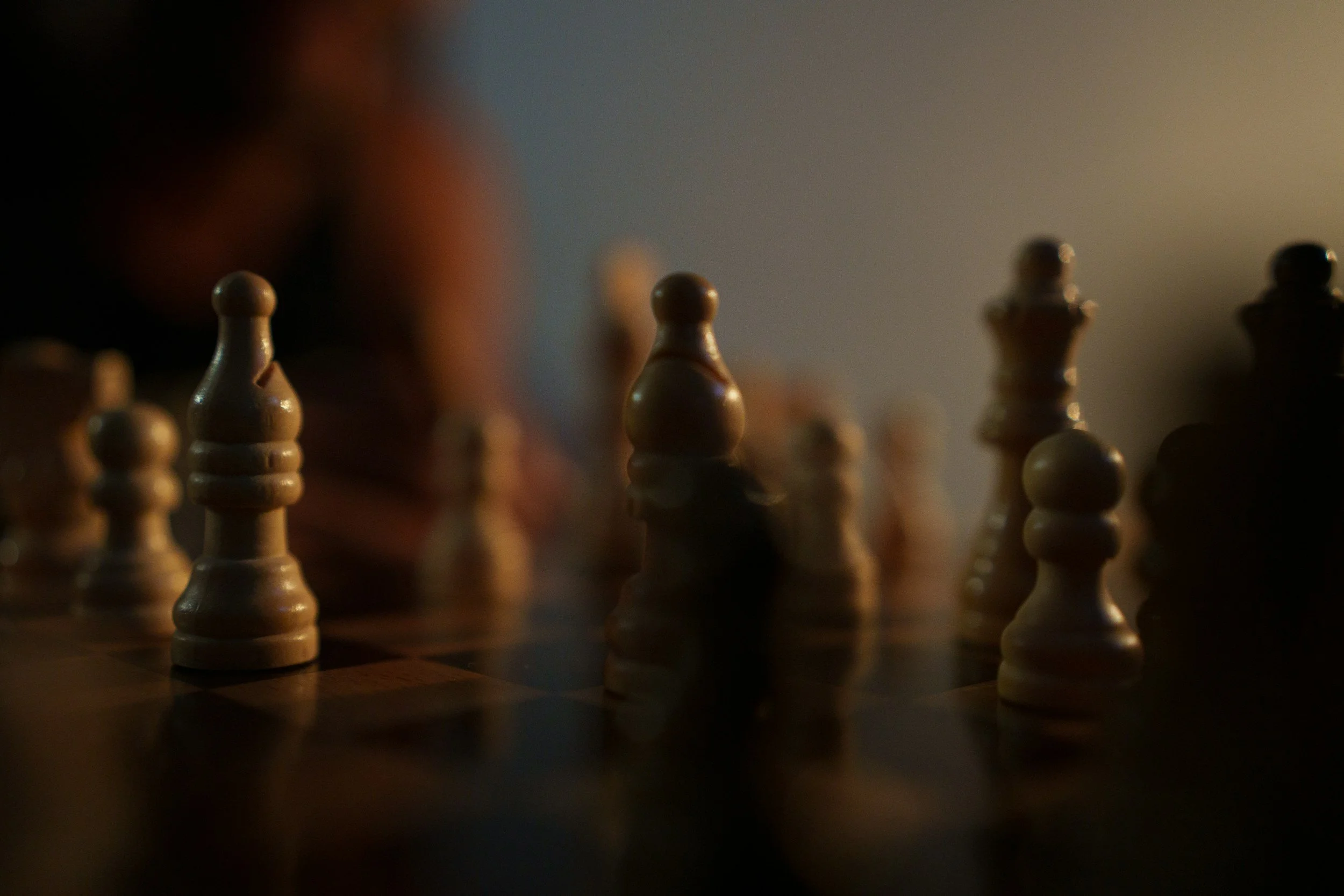 Close-up of a chessboard with wooden chess pieces, with a player visible in the background, softly lit.