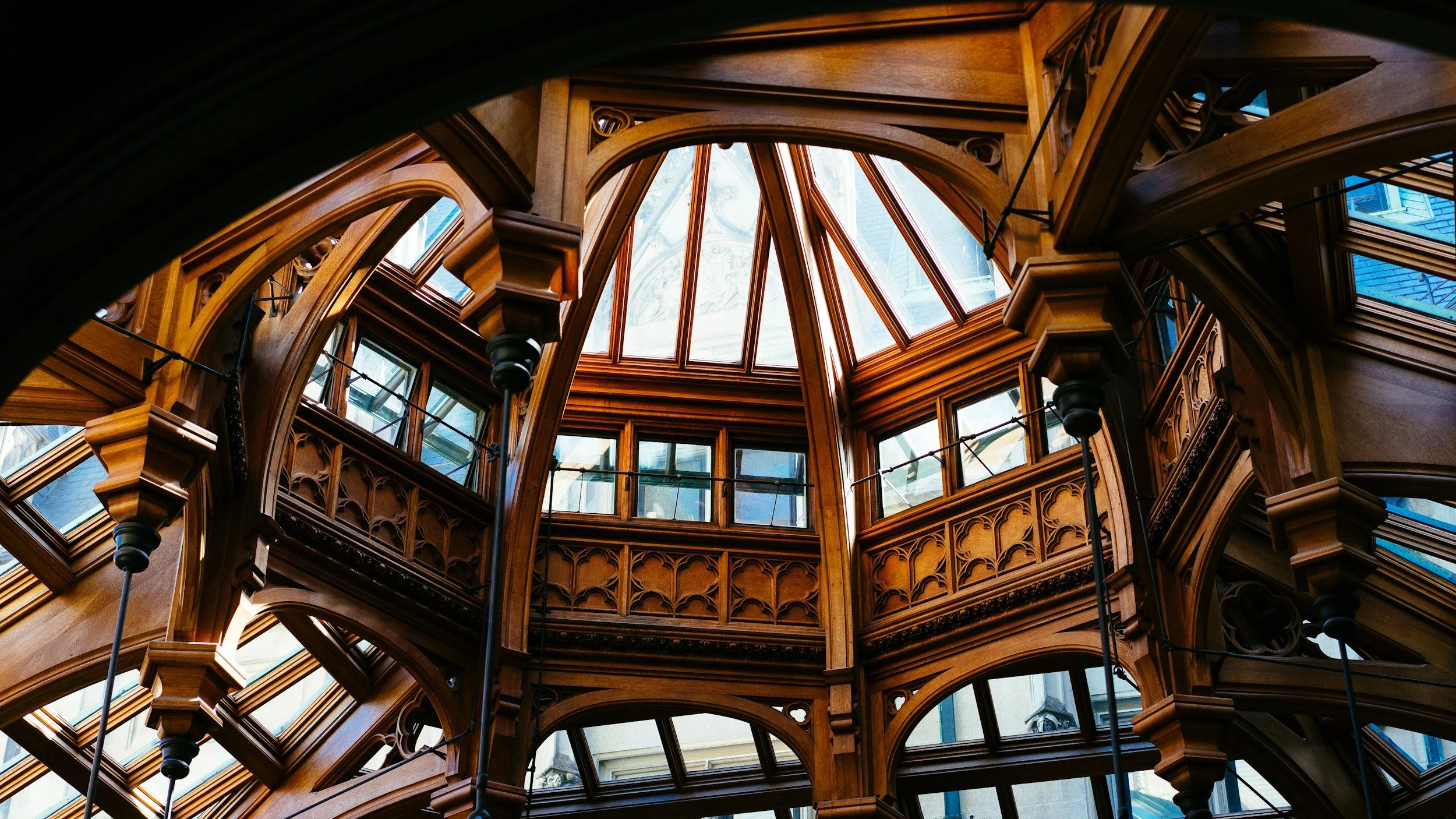 Interior view of a multi-story wooden atrium with intricate woodwork, large windows, and a glass ceiling.