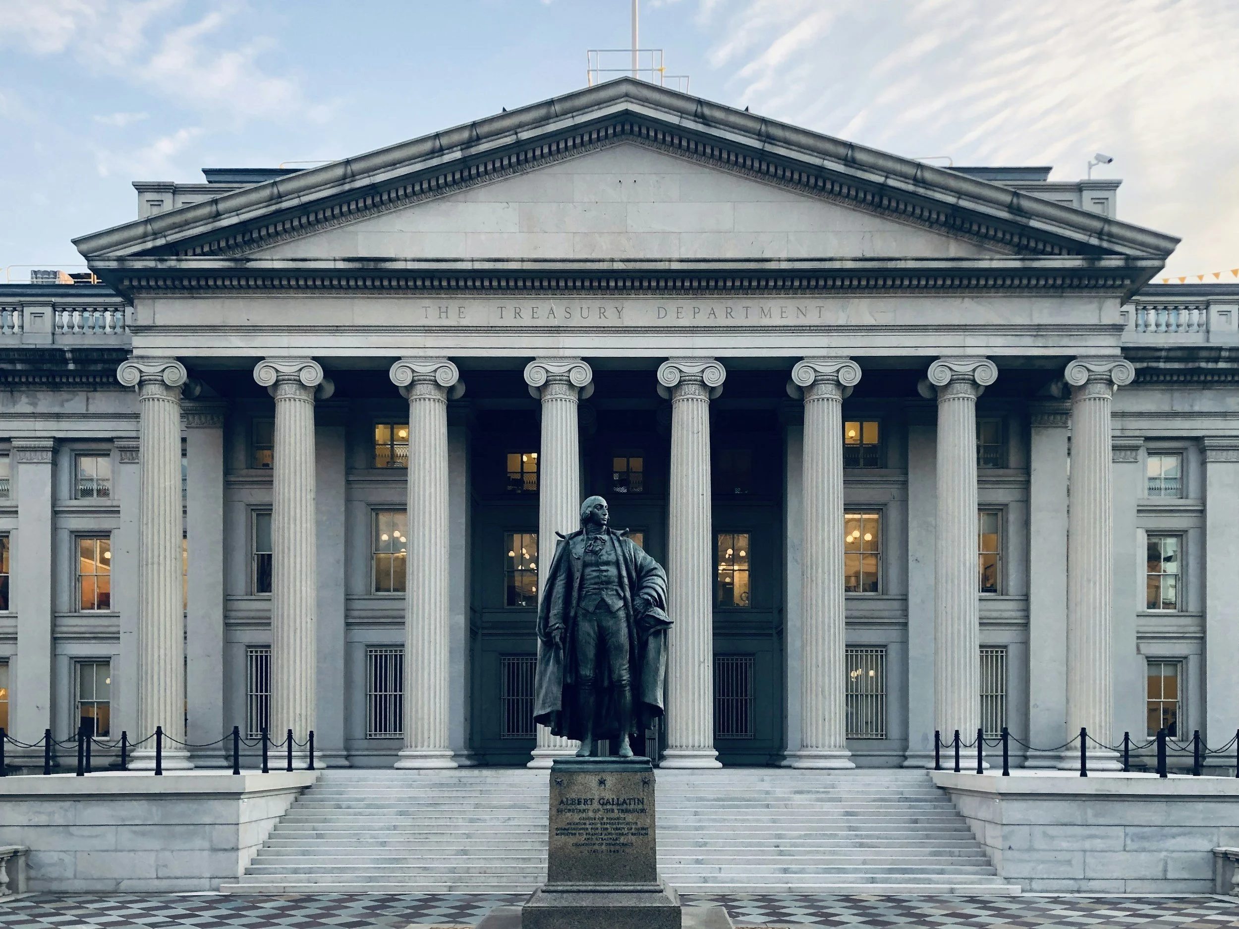 Front view of the Treasury Department building with a statue of Albert Gallatin in front, stairs leading up to the entrance, and a checkered pavement.
