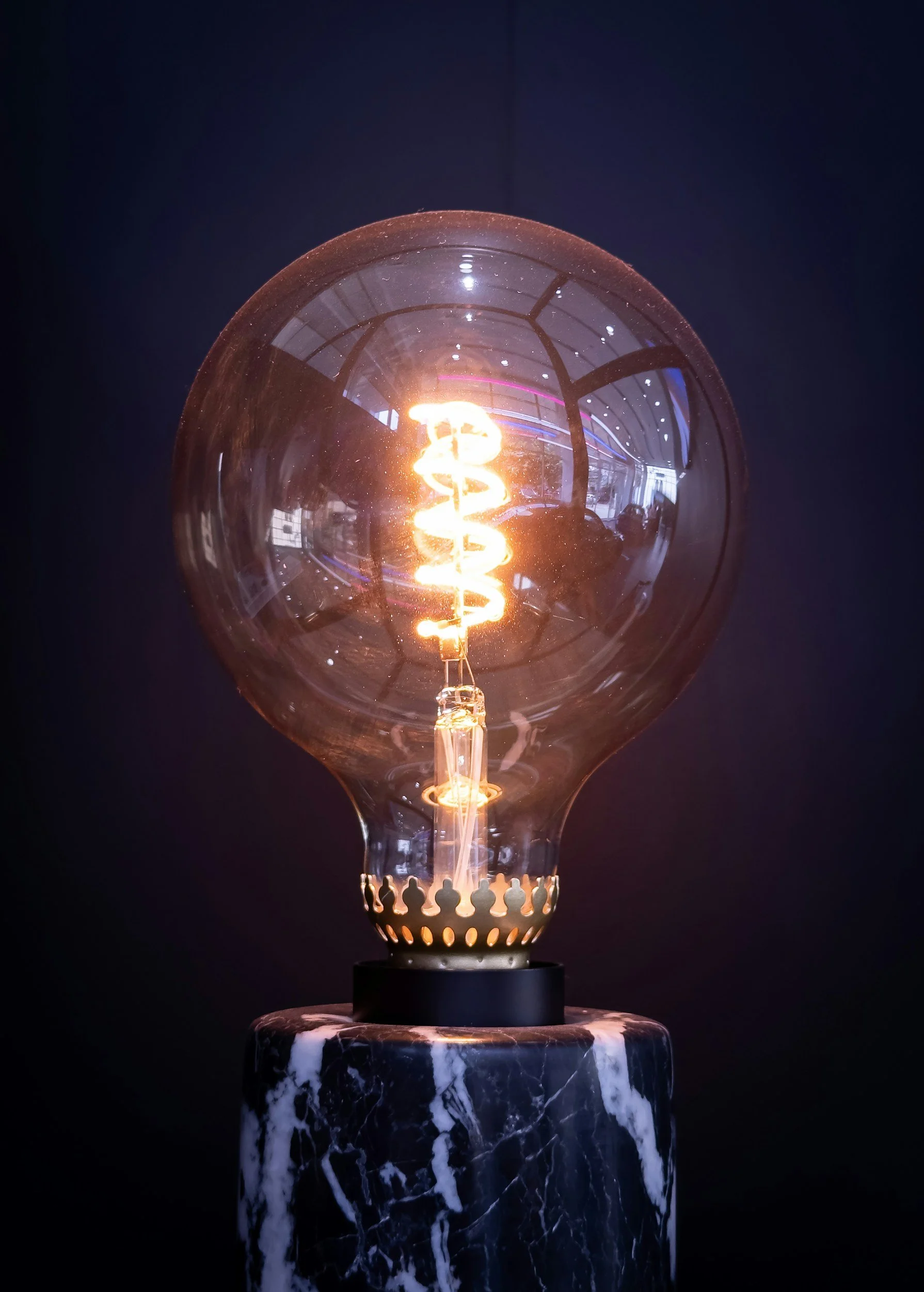 A glowing Edison-style filament light bulb on a marble base, with a dark background and reflections showing a room and windows.