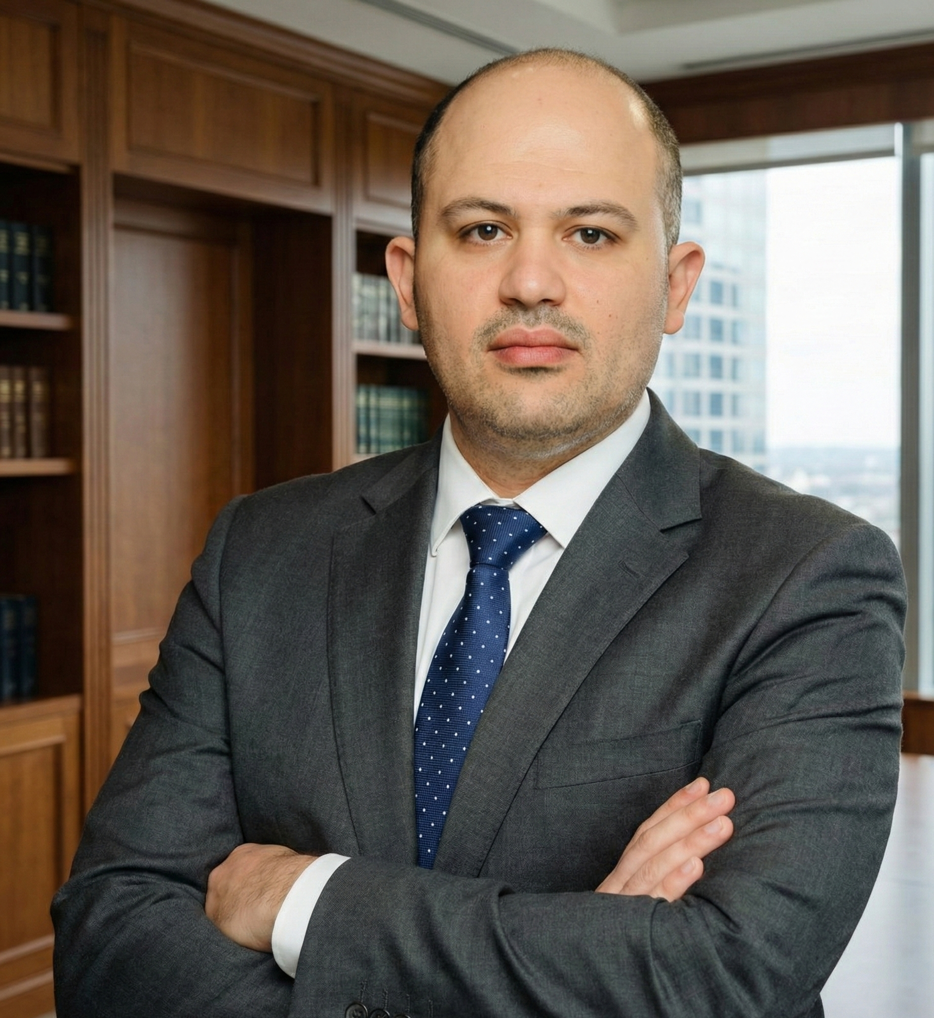 A man in a business suit with folded arms standing in an office with wooden bookshelves and a large window.