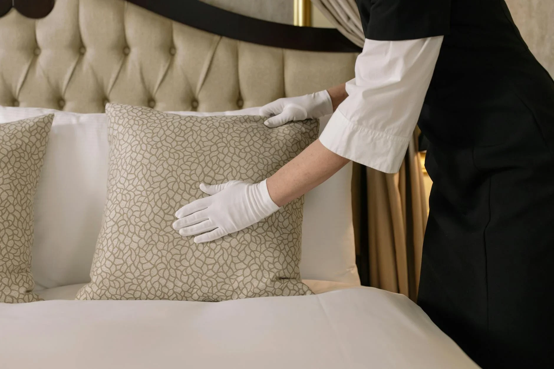 En person som bär vit handskar viks in ett dekorativt kuddfodral på ett lyxigt sängklädesbord i ett hotellrum.
