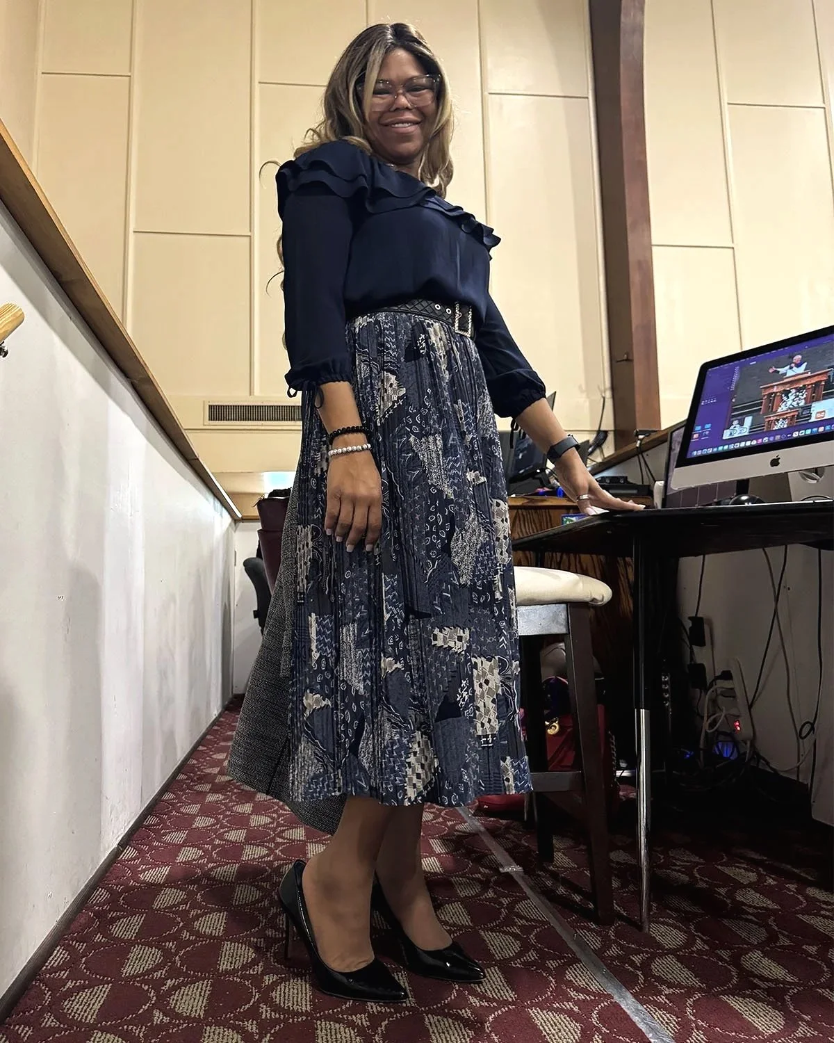 A woman standing beside a desk with a computer, smiling, dressed in a navy blue blouse, a patterned chiffon midi skirt, and high heels.