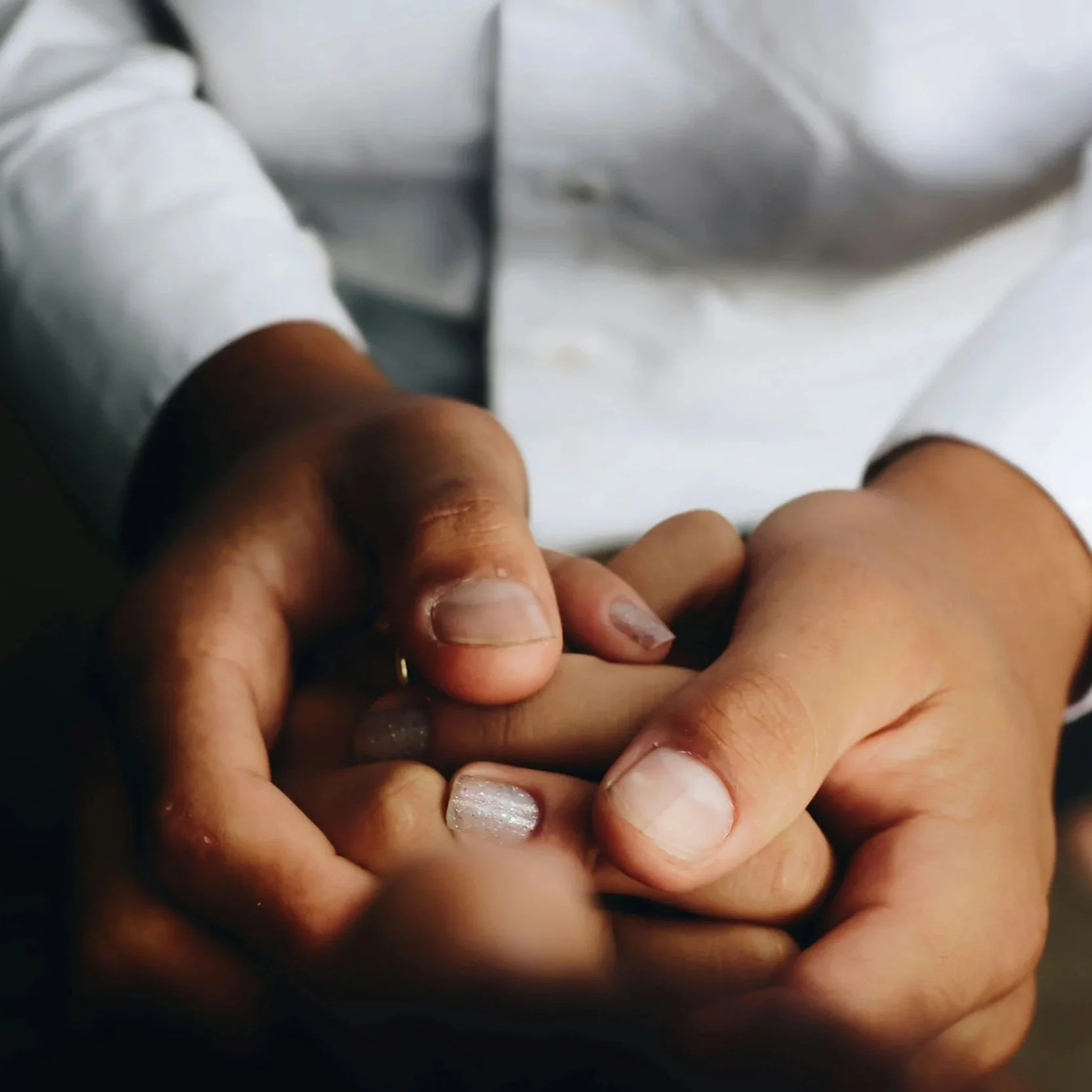 Close-up of two people holding hands gently, one with darker skin and the other with lighter skin, both having natural nails, with a white shirt sleeve visible.