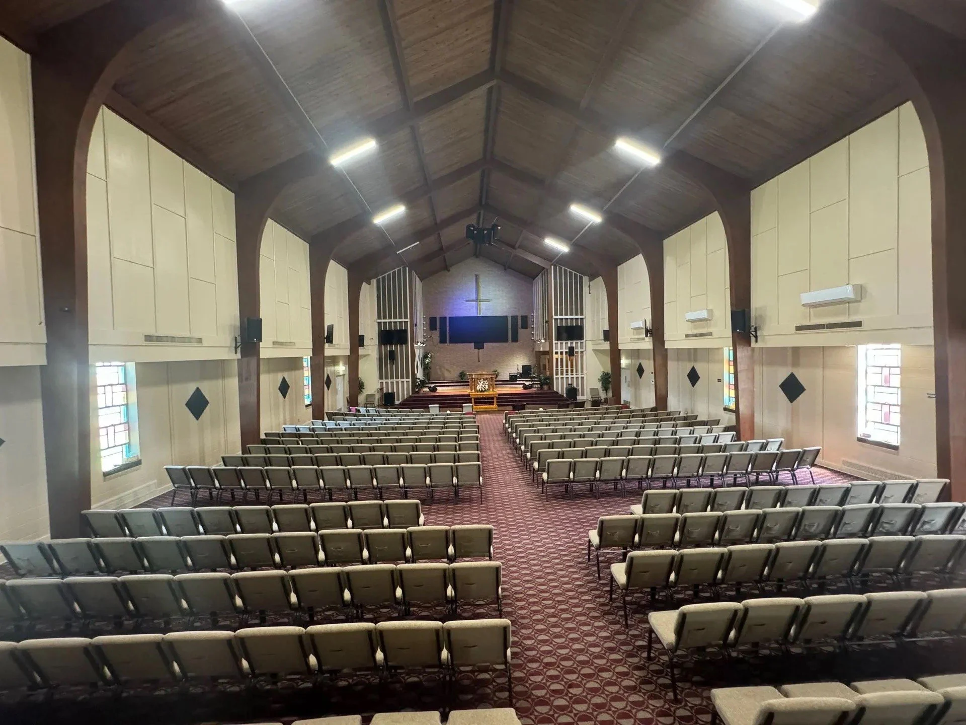 Interior of a church with multiple rows of beige padded chairs arranged facing a stage with a wooden altar, cross, and stained glass windows on the sides.