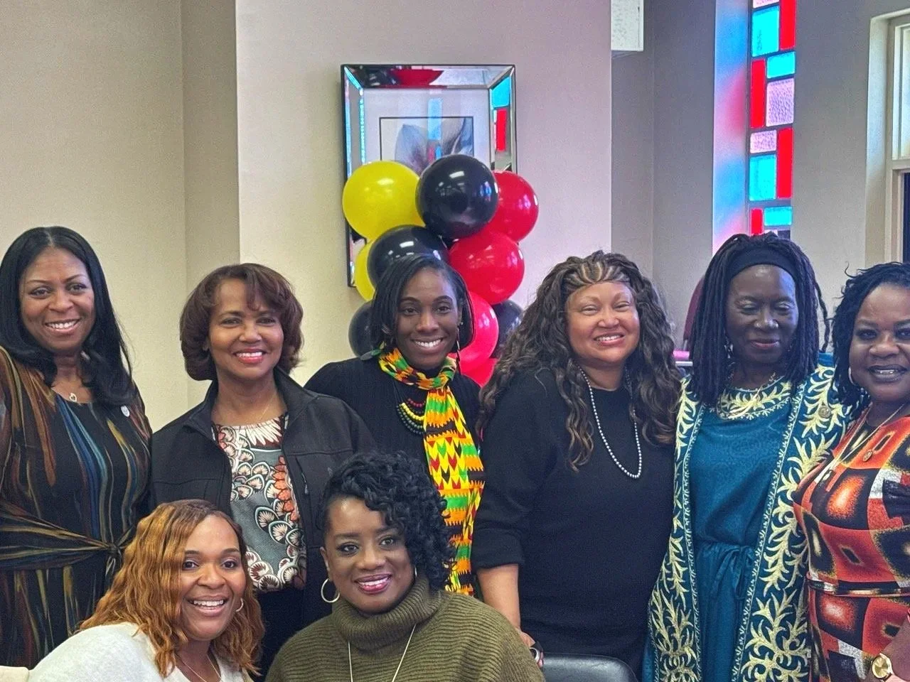 Group of eight women smiling at an indoor event, with colorful balloons and stained glass windows in the background.