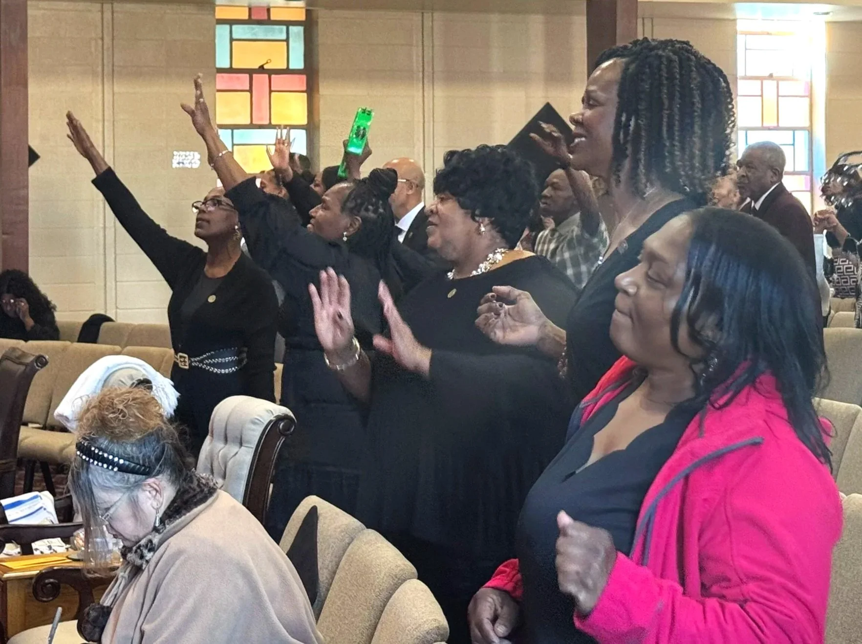 Group of people standing inside a church, with some raising their hands in worship and others with eyes closed in prayer, stained glass windows in the background.