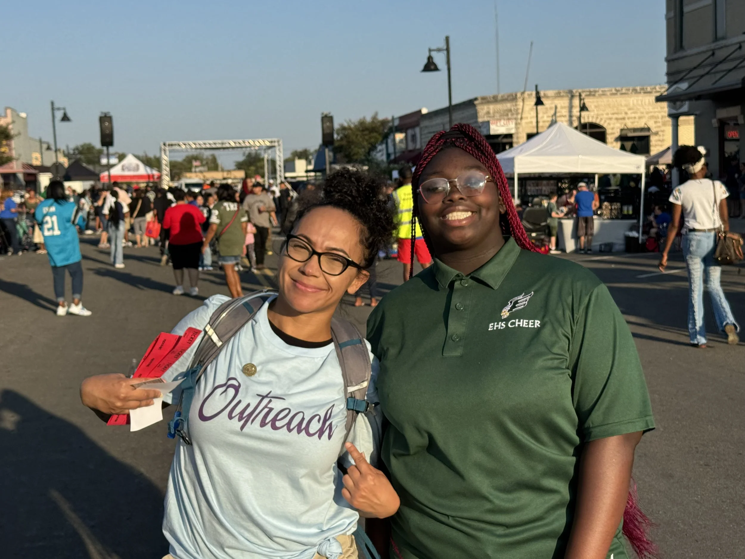 Two smiling young women standing together at an outdoor event, one wearing a light blue Outreach shirt and the other a green EHS Cheer shirt, with a busy street scene and crowd in the background.