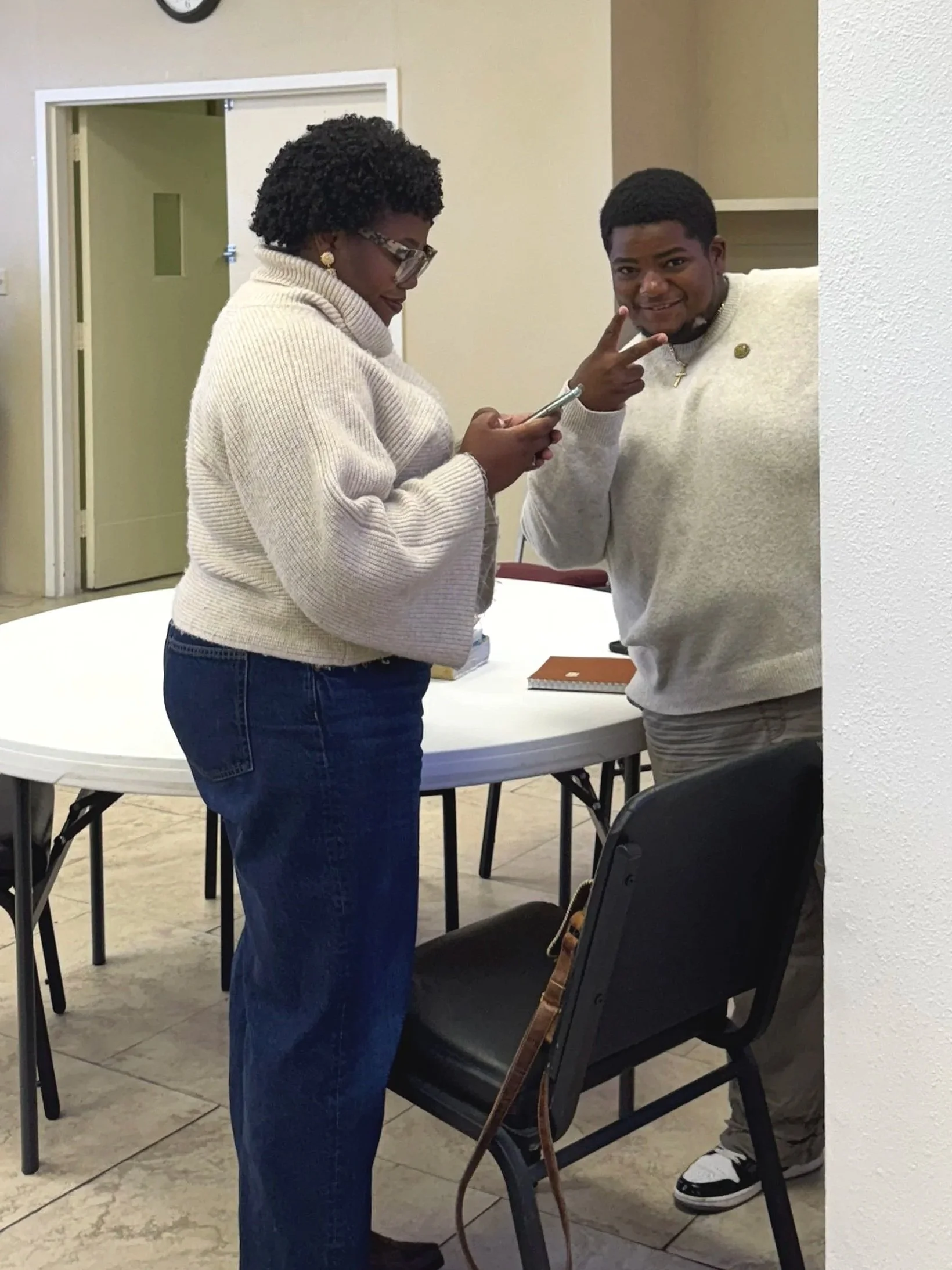 A woman with glasses and earrings looking at her phone, and a young man smiling and making a peace sign near a table in a room with beige walls.