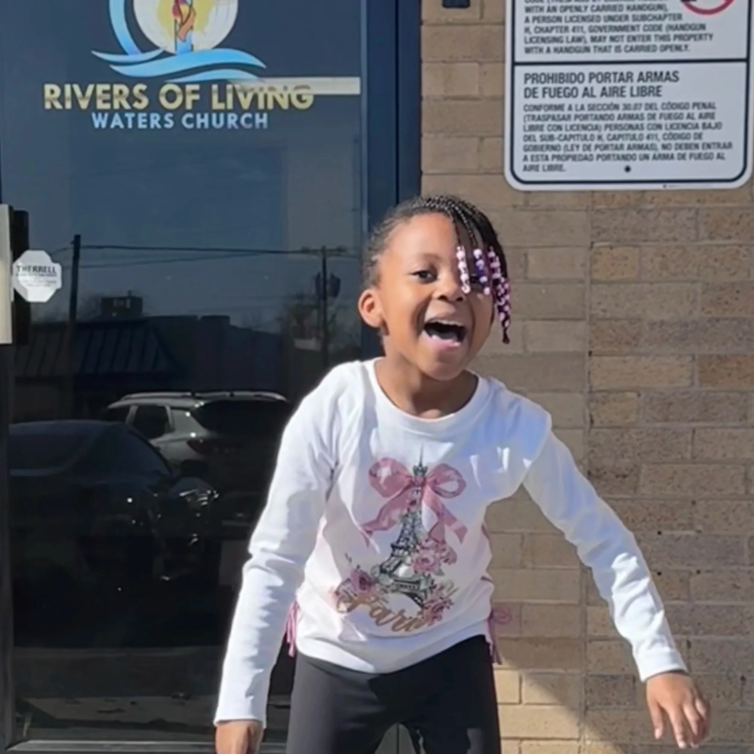 A young girl with braided hair and purple beads, smiling and laughing outdoors in front of a brick wall and a glass door with a church sign.