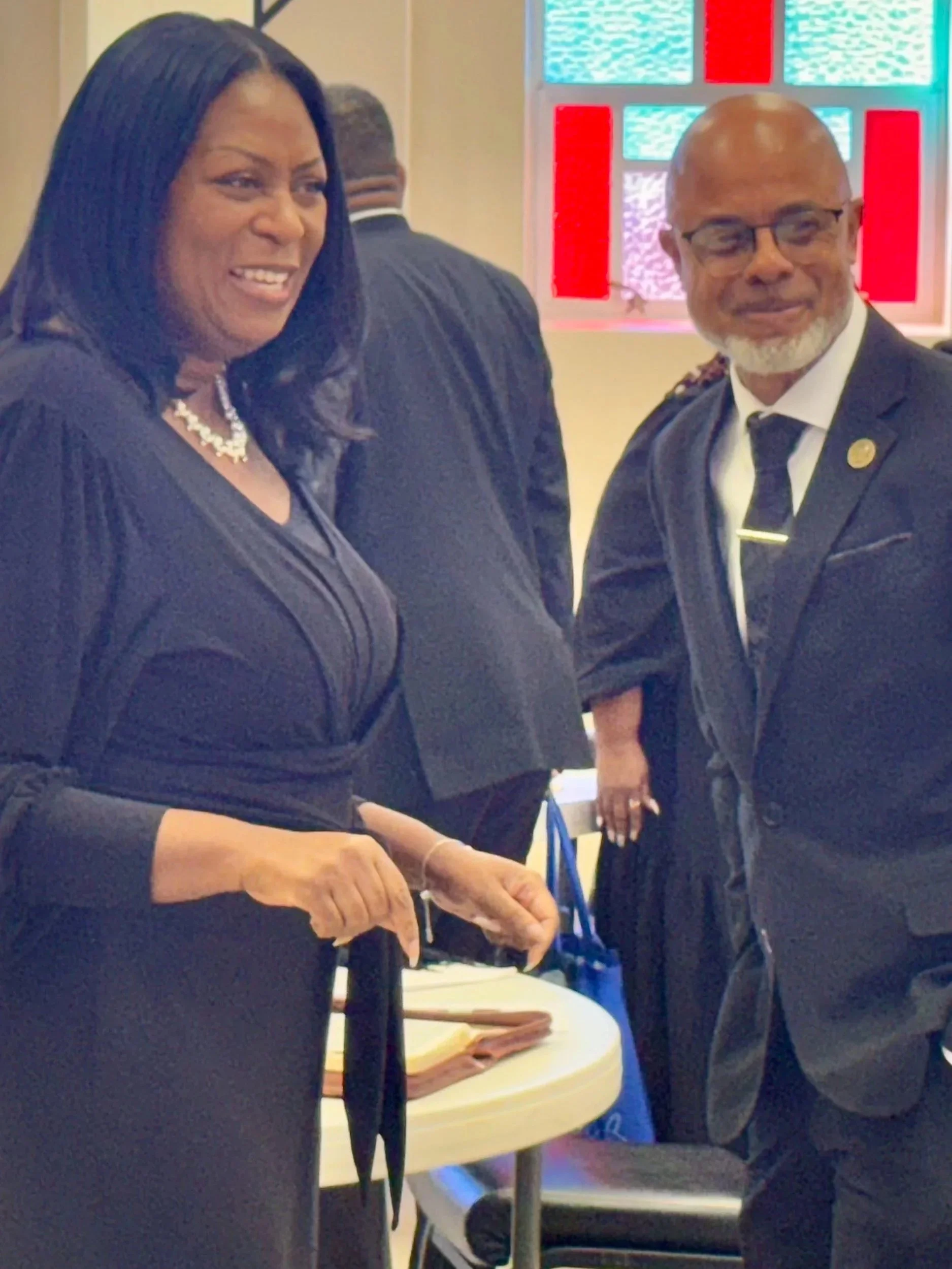 Two professionals, a woman and a man, dressed in formal business attire, are standing and smiling at a professional event or conference near a table with documents, with stained glass windows in the background.