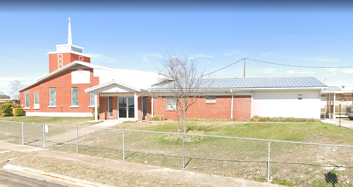 A small church building with red brick exterior, white steeple, and a surrounding chain-link fence, located on a grassy lawn with a sidewalk in front.