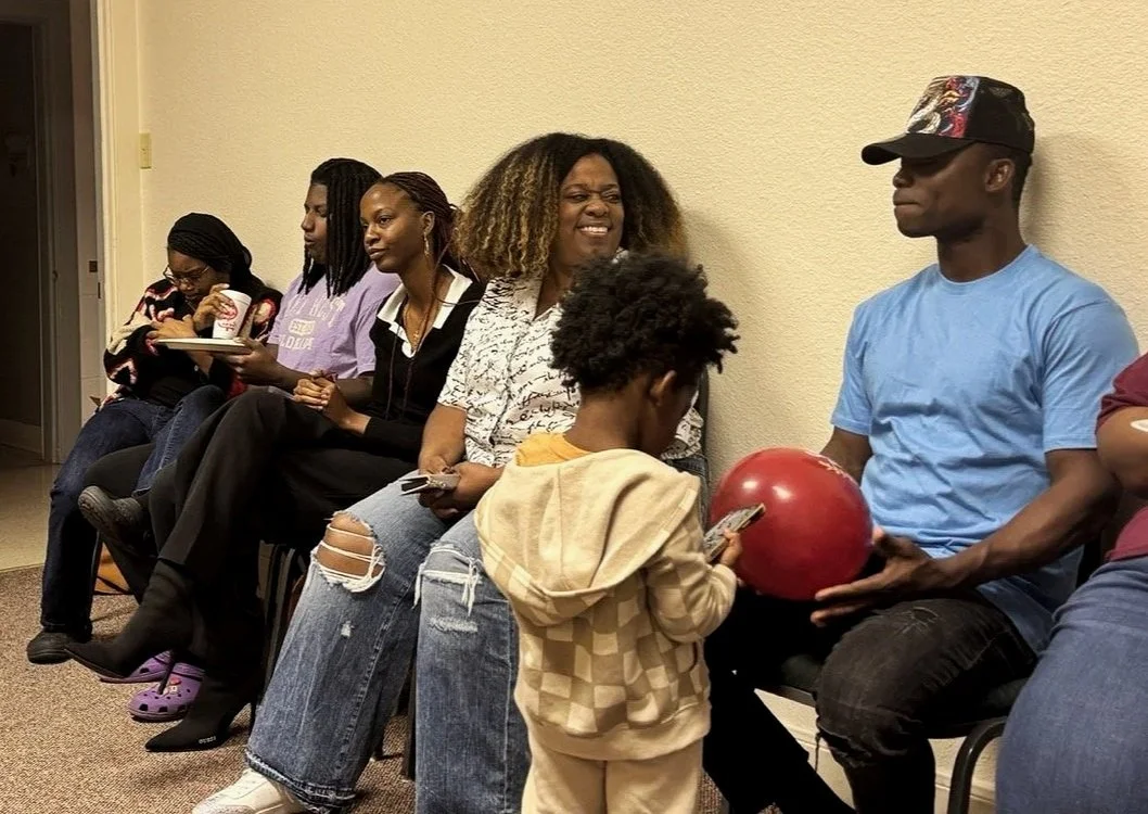 People sitting in a line against a beige wall, with a woman holding a red balloon and a young boy using a smartphone.