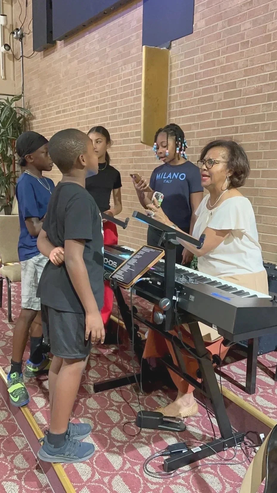 A woman playing a keyboard surrounded by four children, some holding smartphones, in an indoor setting with a brick wall background.