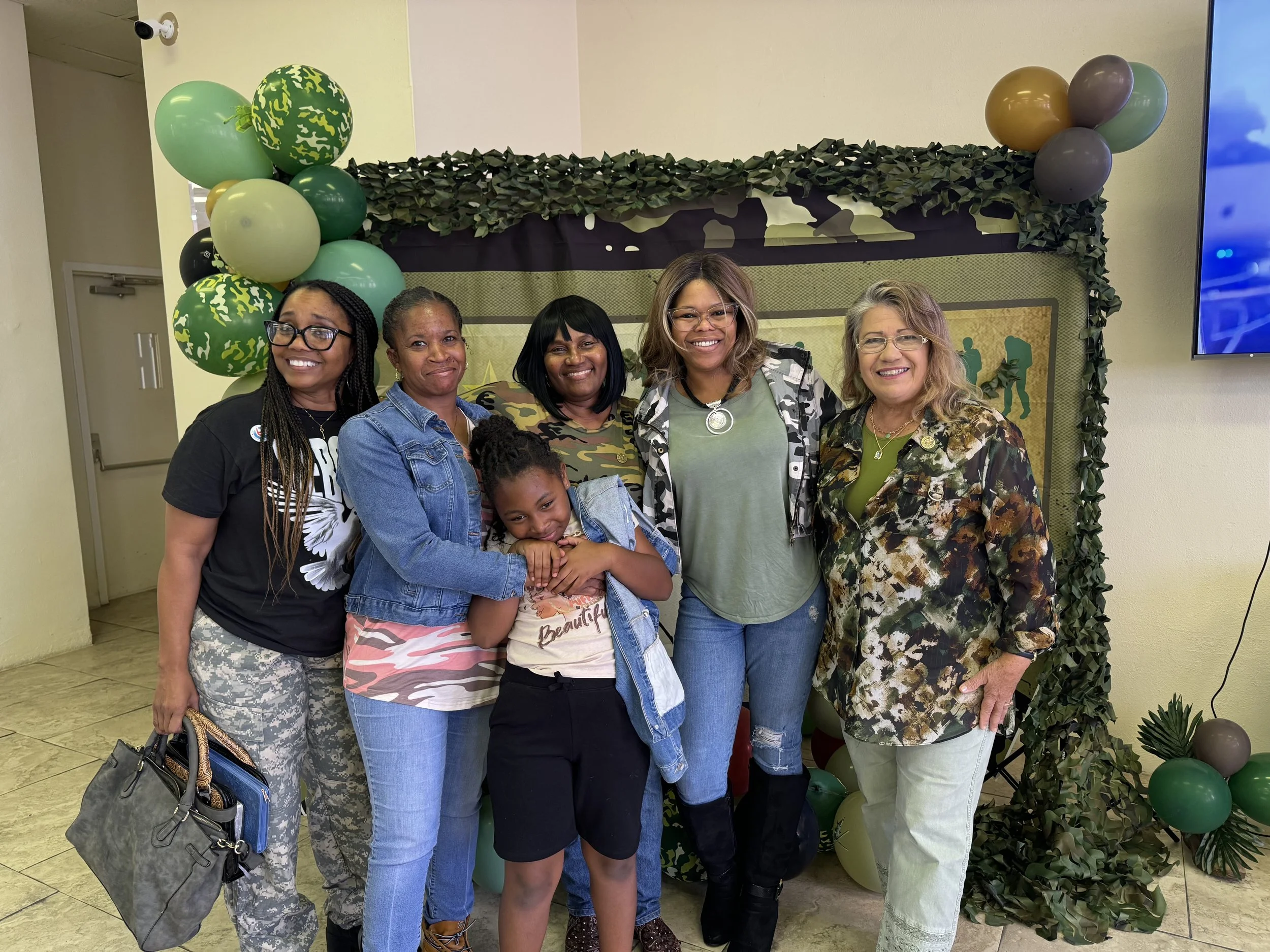 Six women and one girl standing together in front of a decorated backdrop with balloons and greenery, smiling at the camera.