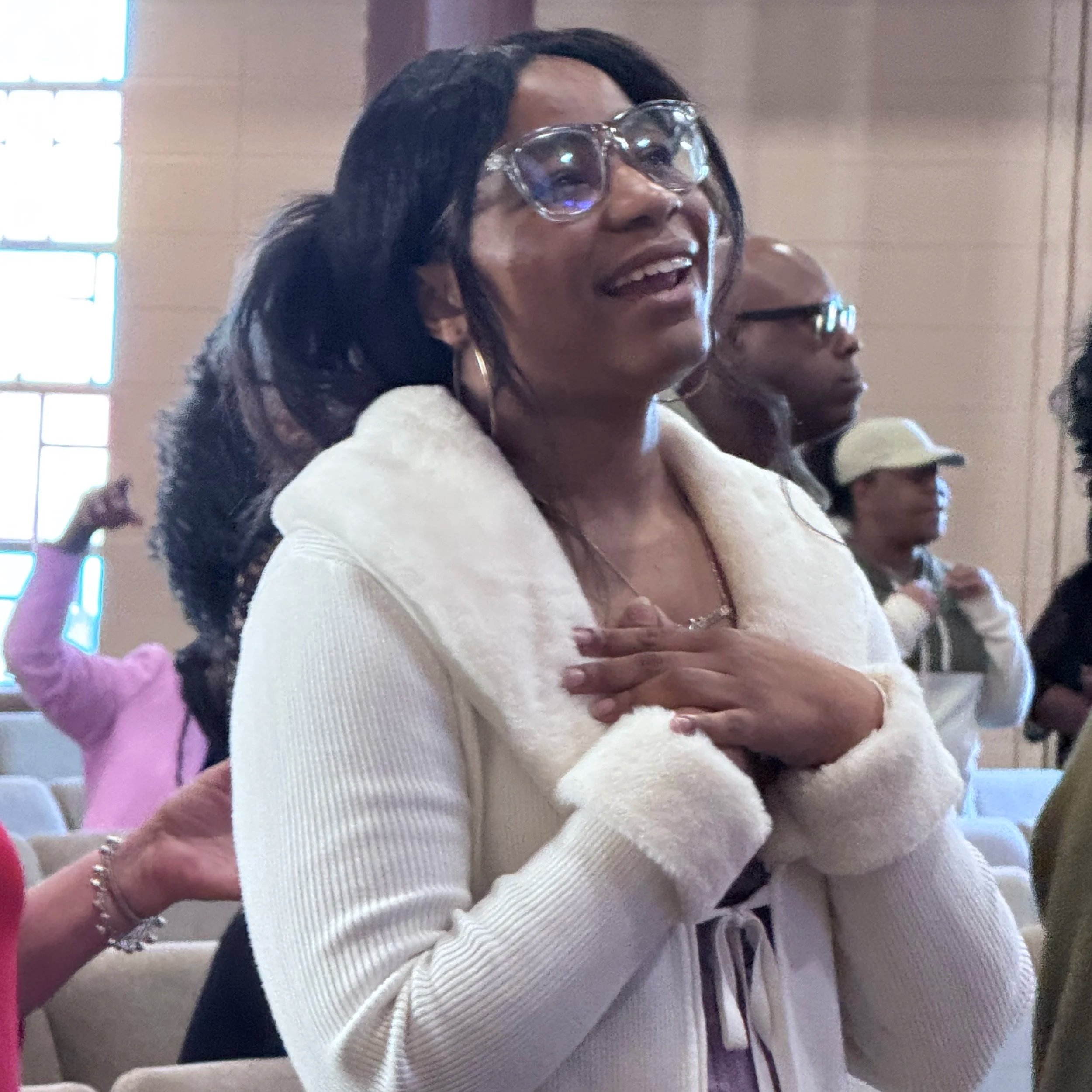 A woman with dark hair and glasses standing indoors with her hand over her chest, smiling and looking upward. There are other people in the background.